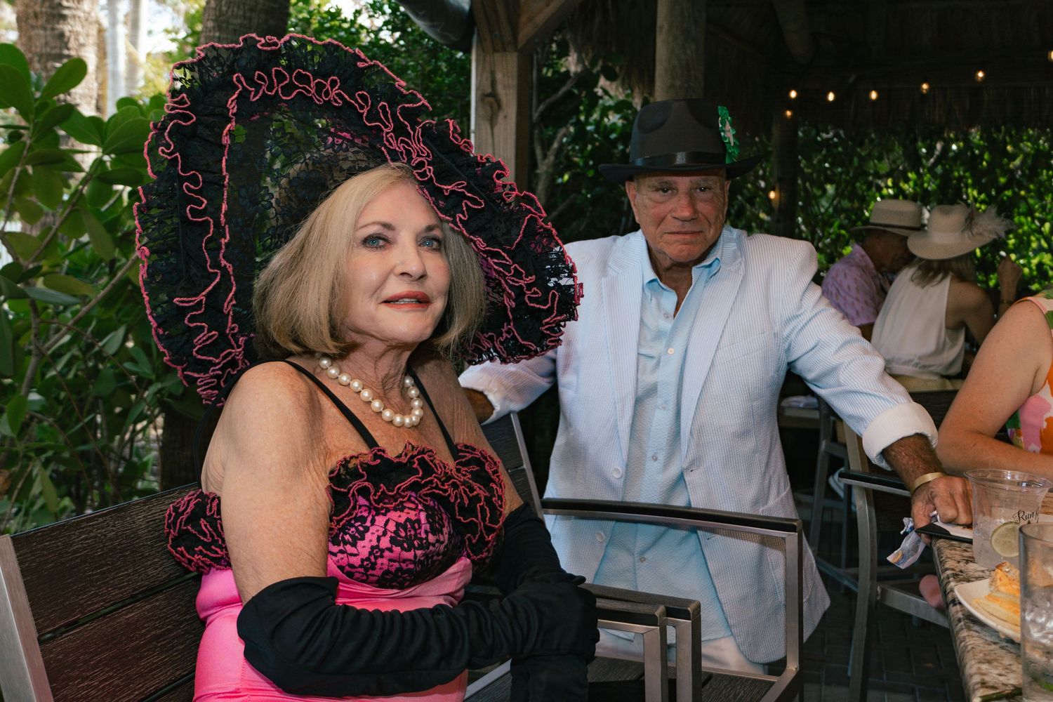 Woman in pink dress and black feathered hat seated with man in white jacket at outdoor garden event.