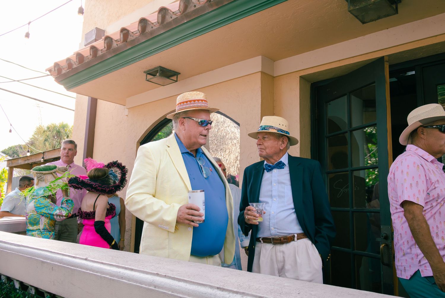 Two men in summer suits and hats socializing on a patio at a Kentucky Derby-style outdoor party.