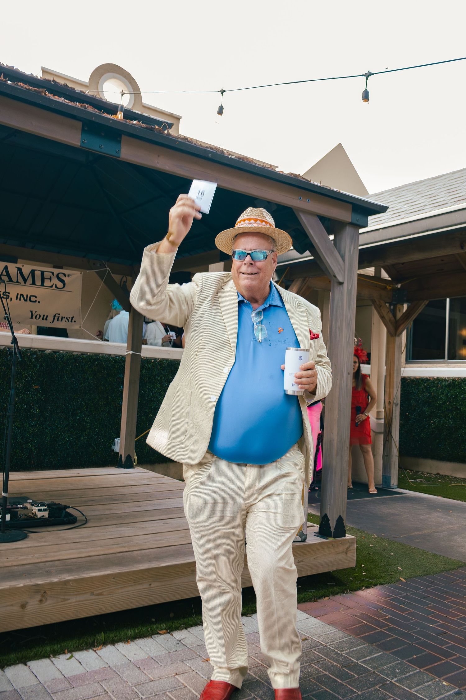 Festive man in straw hat and blue polo raising card while holding drink at outdoor event.