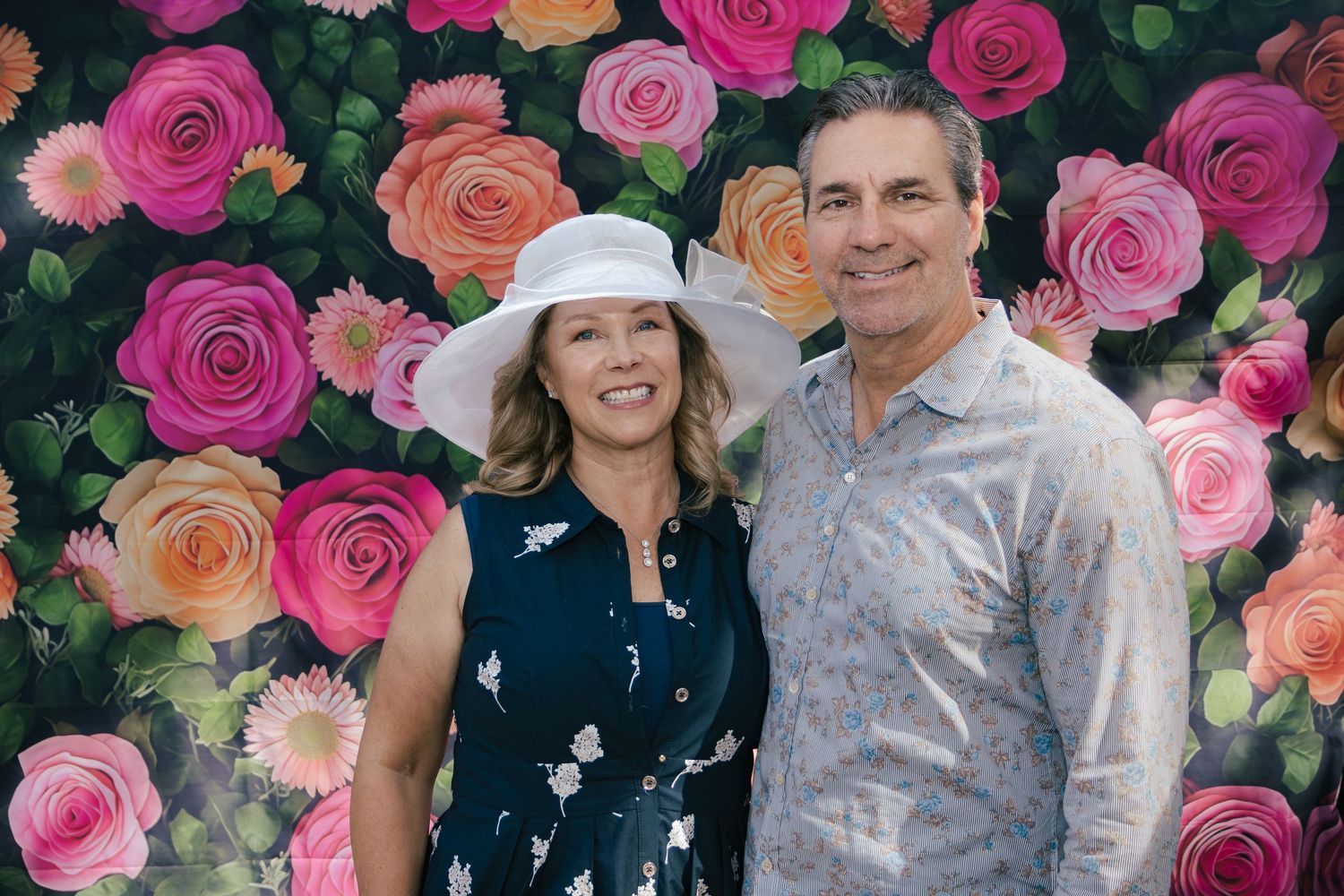 Smiling couple posing in front of a colorful floral rose backdrop at a Kentucky Derby themed event.