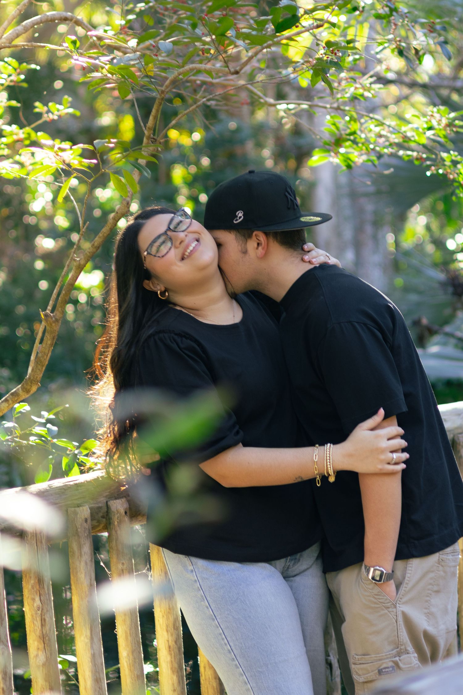 Young man kissing woman on cheek while embracing outdoors near wooden fence with green foliage background.