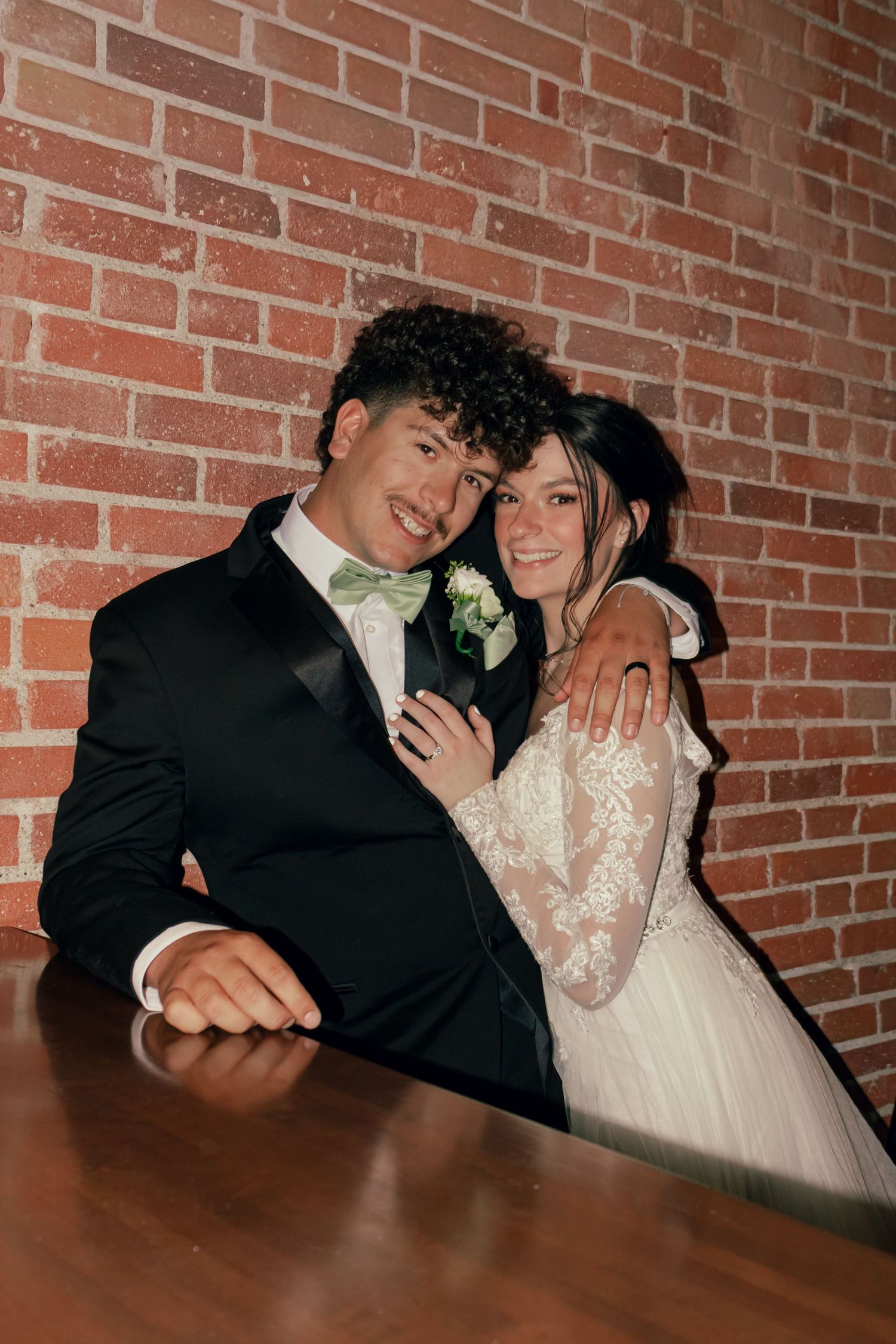 Smiling newlyweds pose at a wooden table against a brick wall, groom in black tuxedo with green bow tie and bride in lace gown.