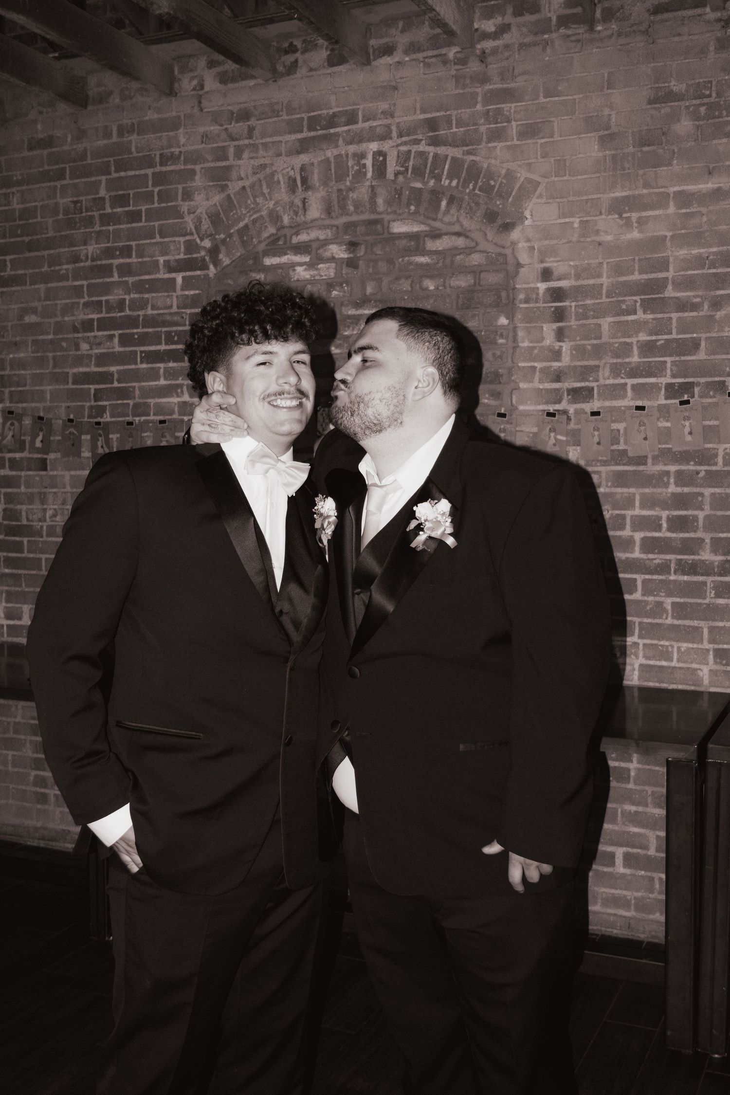 Two groomsmen in black tuxedos share a playful moment against a brick wall.