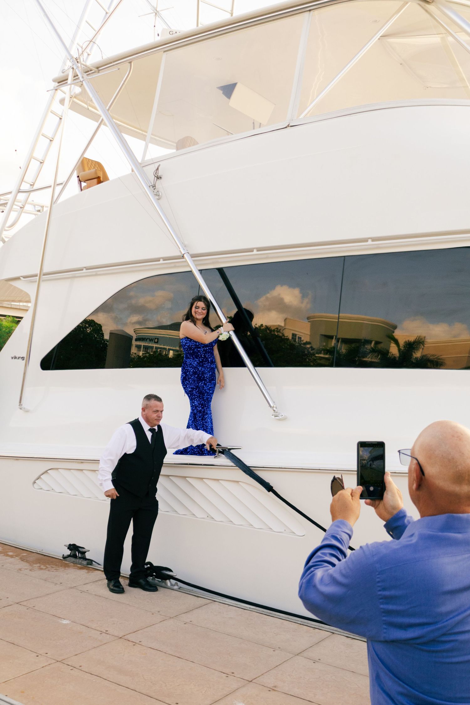 A teenage girl in a blue sequin prom dress poses on a white yacht while a man photographs her.