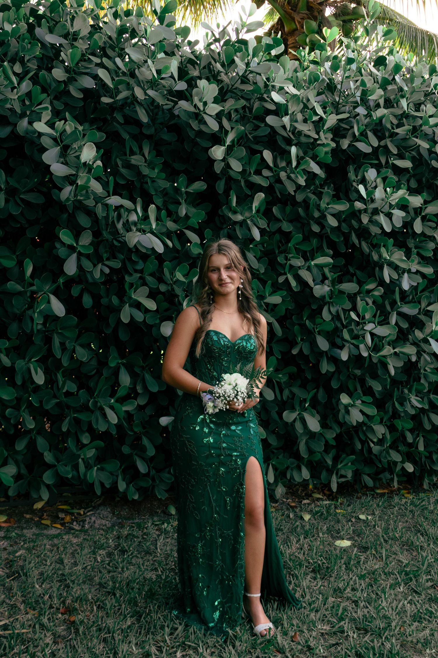 Young woman in an emerald green sequined gown with slit holding white flowers at prom.