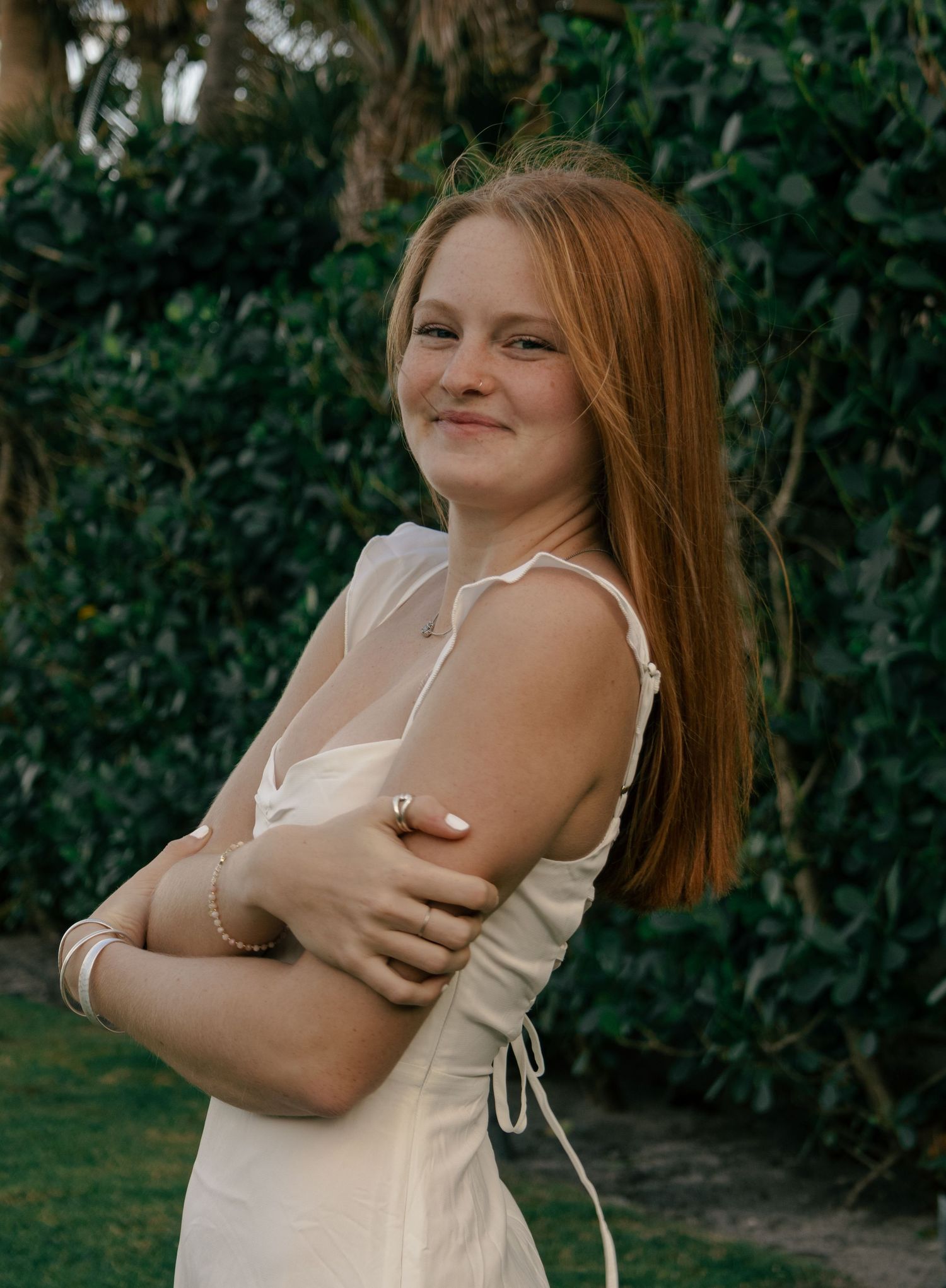 Smiling redhead teenage girl in white dress with arms crossed standing in front of green bushes.