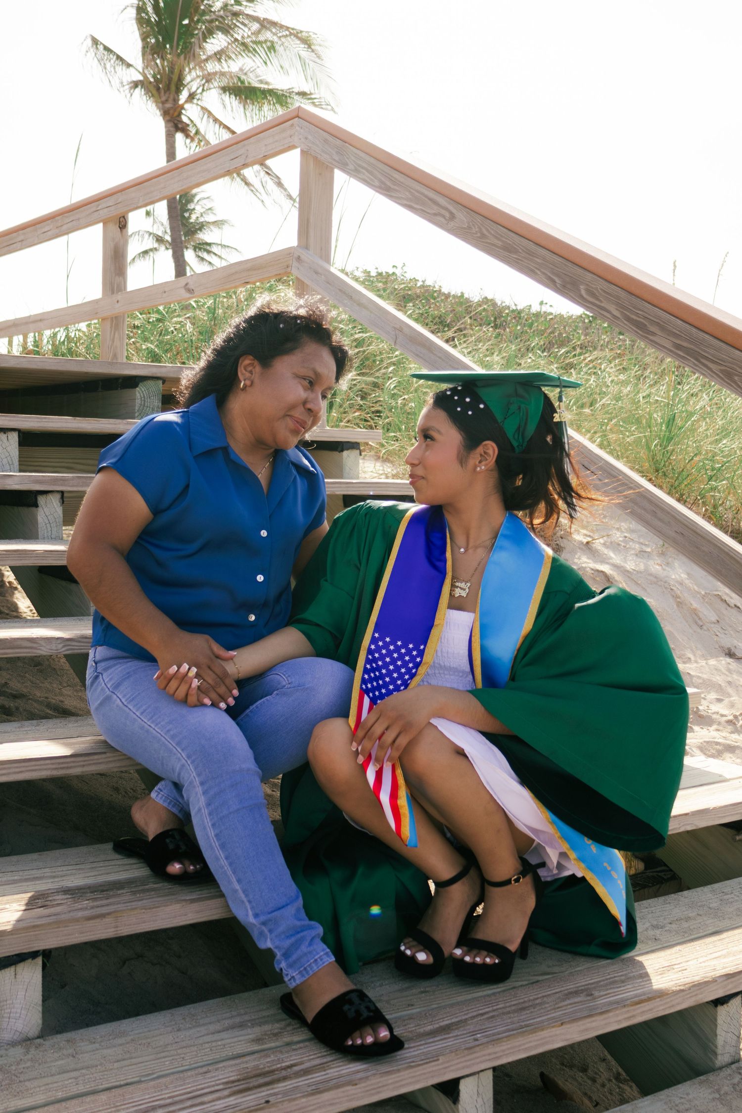 Graduate in green cap and gown sits with family member on wooden beach boardwalk stairs.