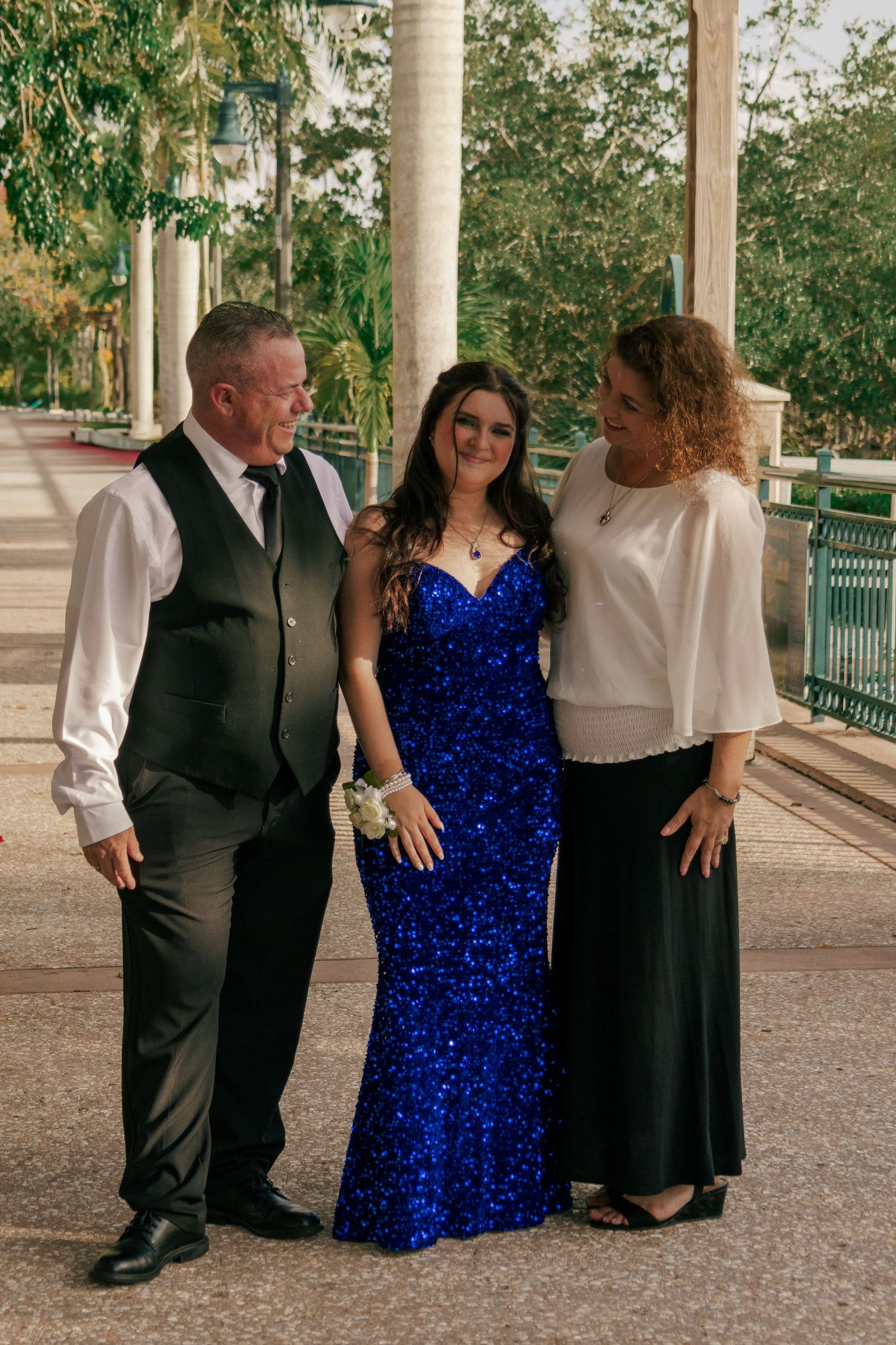 Family portrait under pergola with girl in royal blue sequin gown between parents in formal attire.