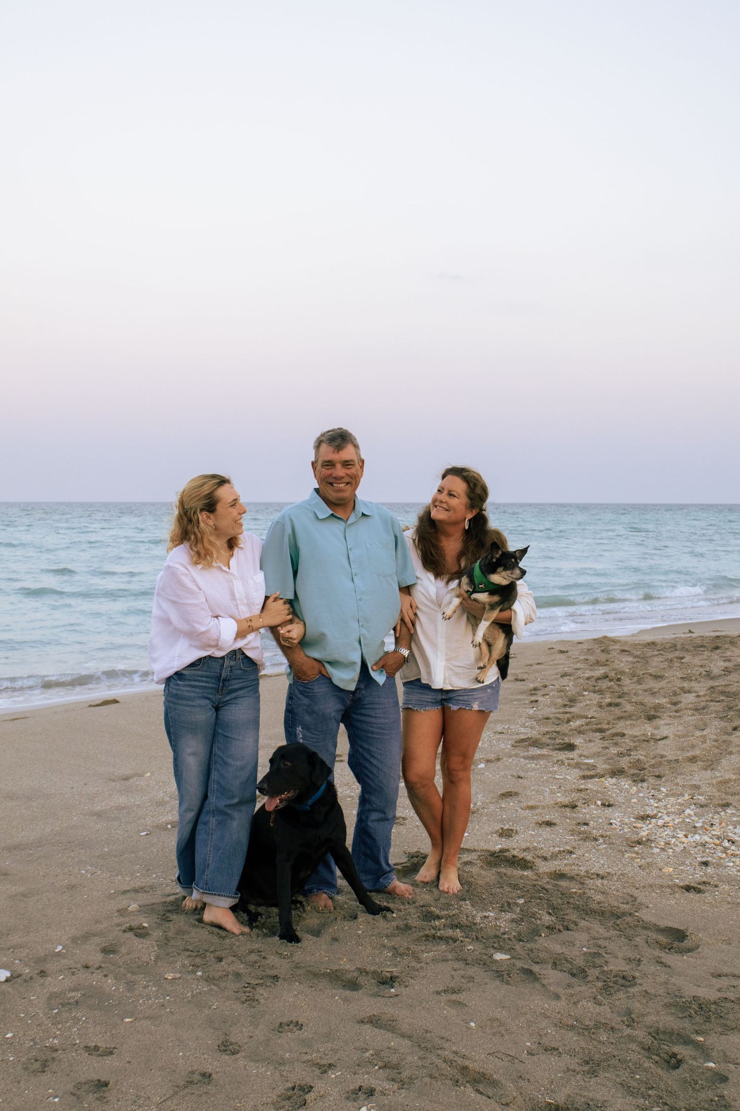 Happy family of three stands with two dogs on beach shore at dusk with calm ocean waves in the background.