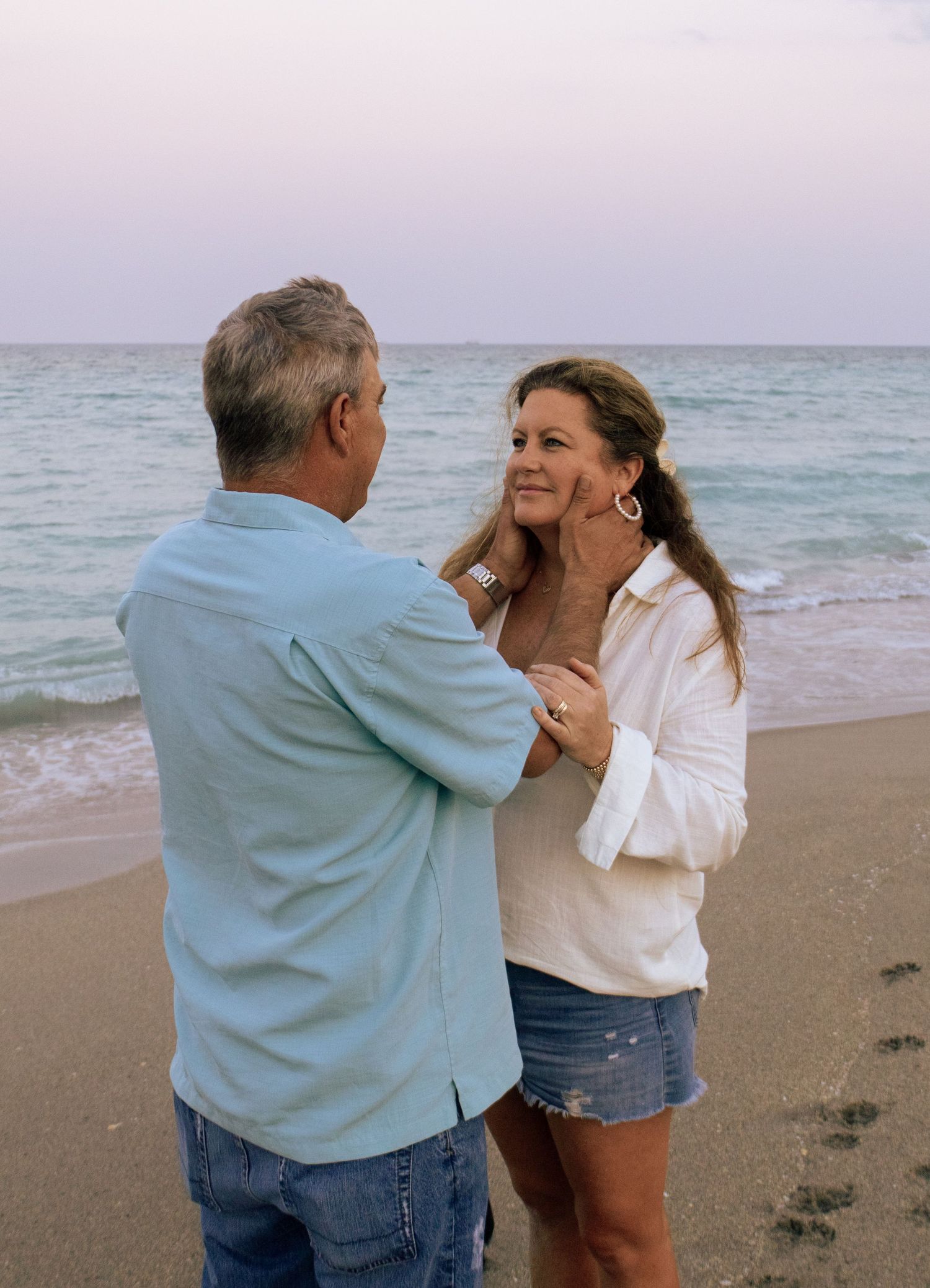 Couple sharing a tender moment on a sandy beach at sunset with ocean waves in the background.