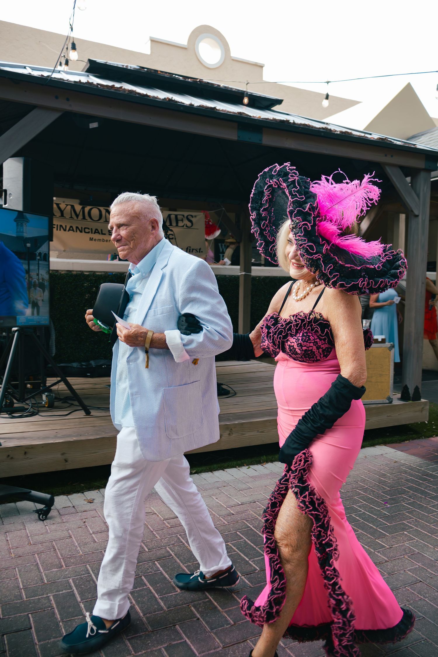 Man in white suit walks alongside woman in pink gown at Kentucky Derby party.