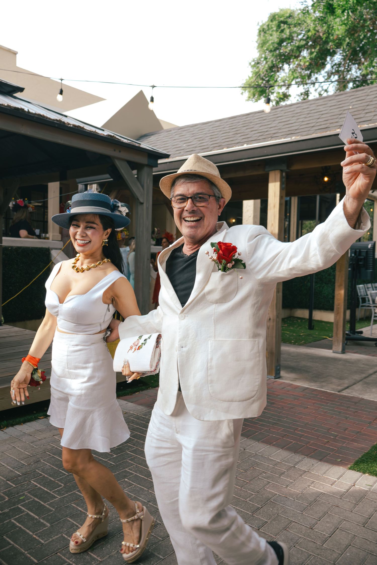 Couple in white outfits dancing outdoors, man in straw hat holding phone, woman in blue fascinator at garden party.