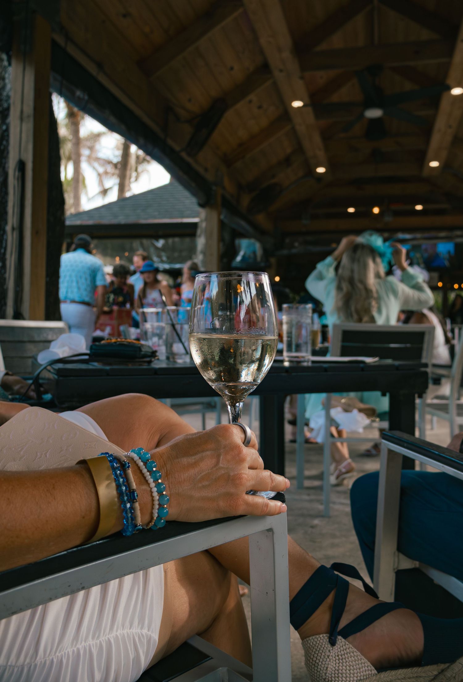 Woman relaxing at outdoor bar holding white wine glass, wearing blue beaded bracelets, with crowd in background.