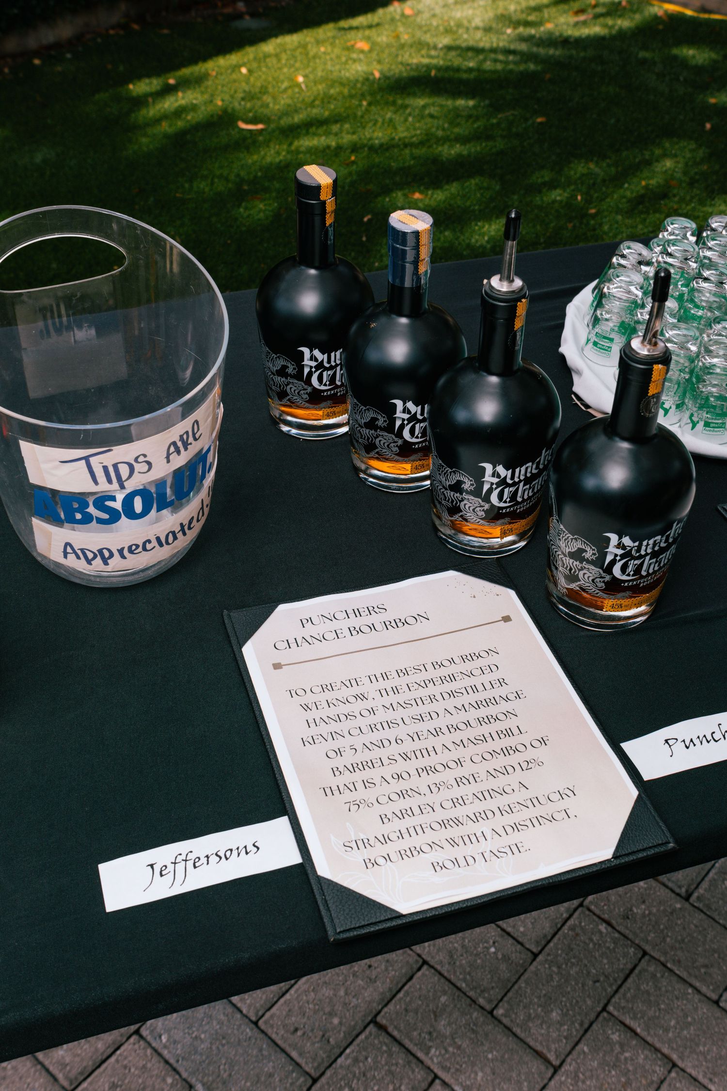 Punchers Chance Bourbon bottles displayed on a black table with a tips jar and cocktail menu at an outdoor bar.