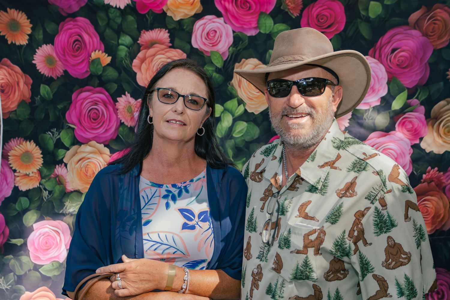 Man and woman smiling together in front of colorful flower wall backdrop at outdoor event.
