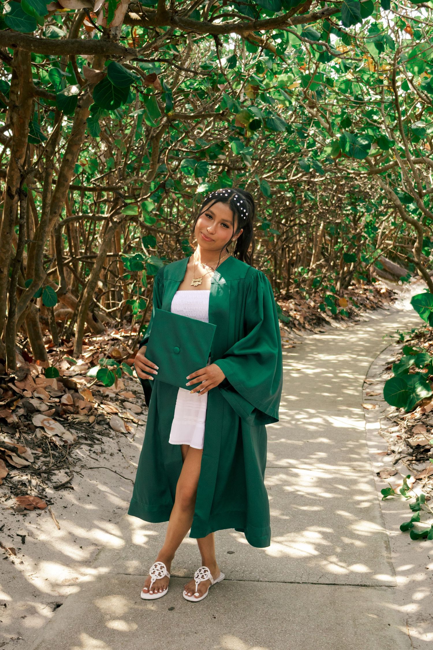 Smiling graduate posing in green cap and gown on shaded sandy pathway surrounded by lush green foliage.
