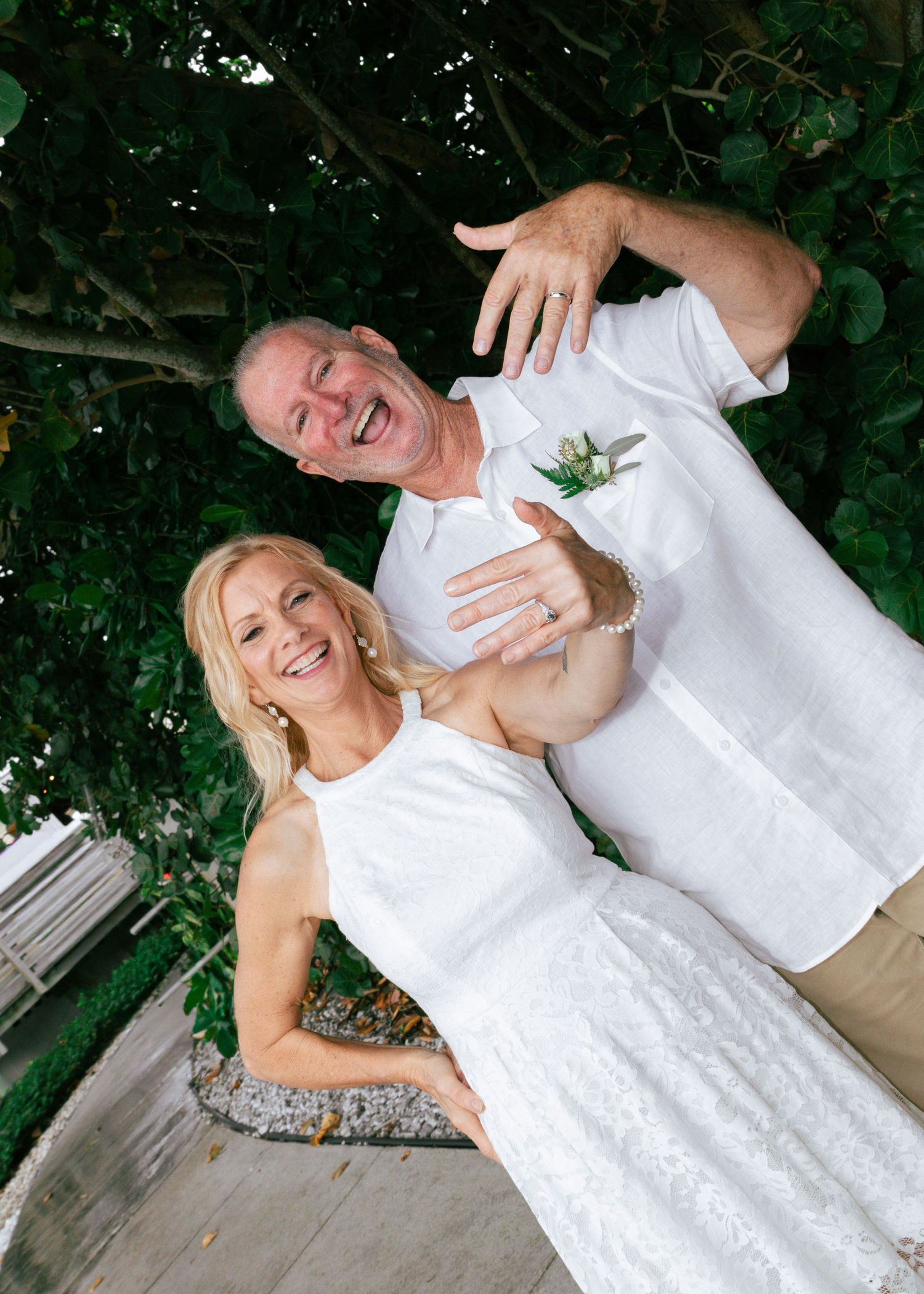 Happy couple in white outfits celebrating their wedding day while showing off rings outdoors.