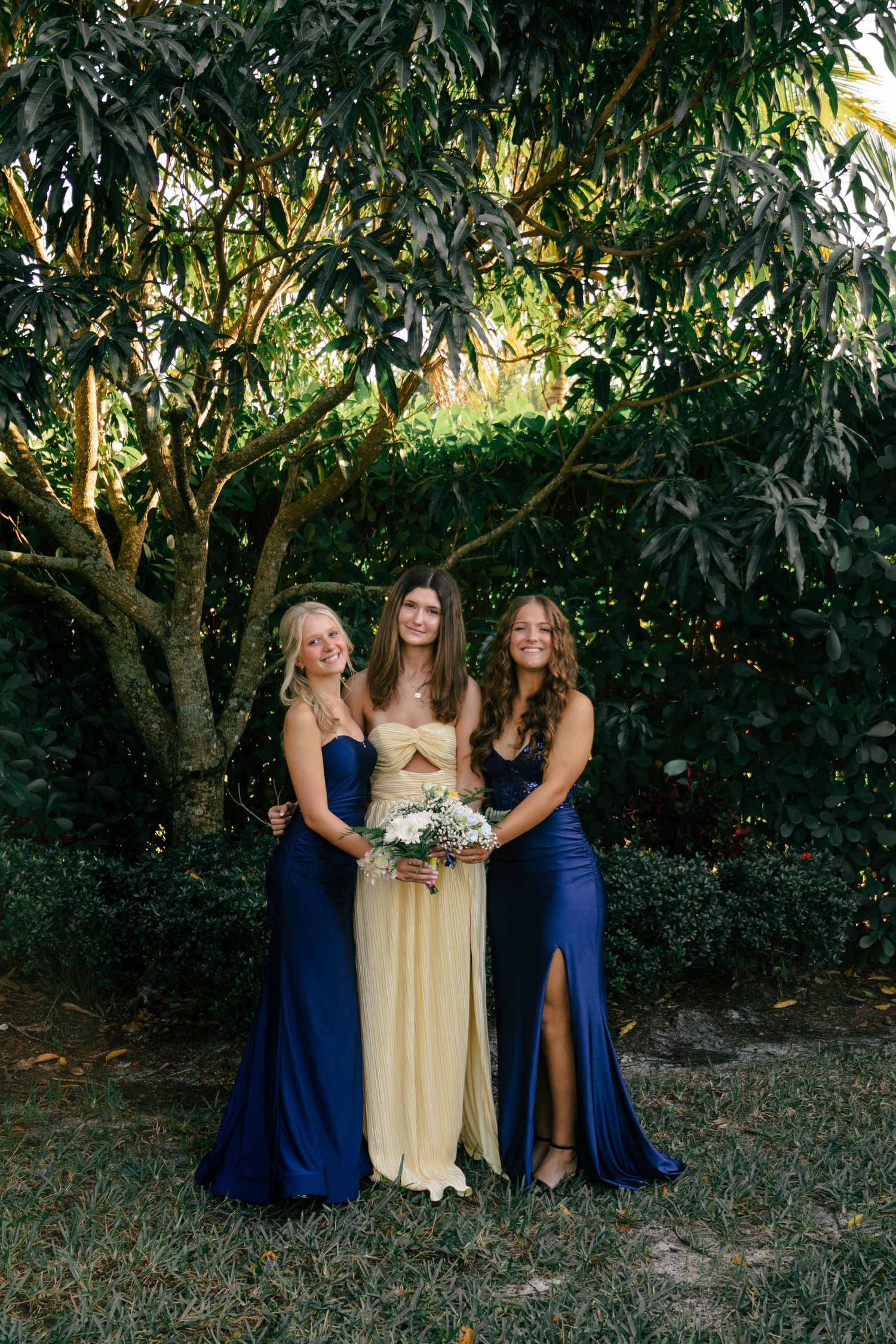 Three young women in navy and yellow prom dresses pose together beneath a lush green tree.
