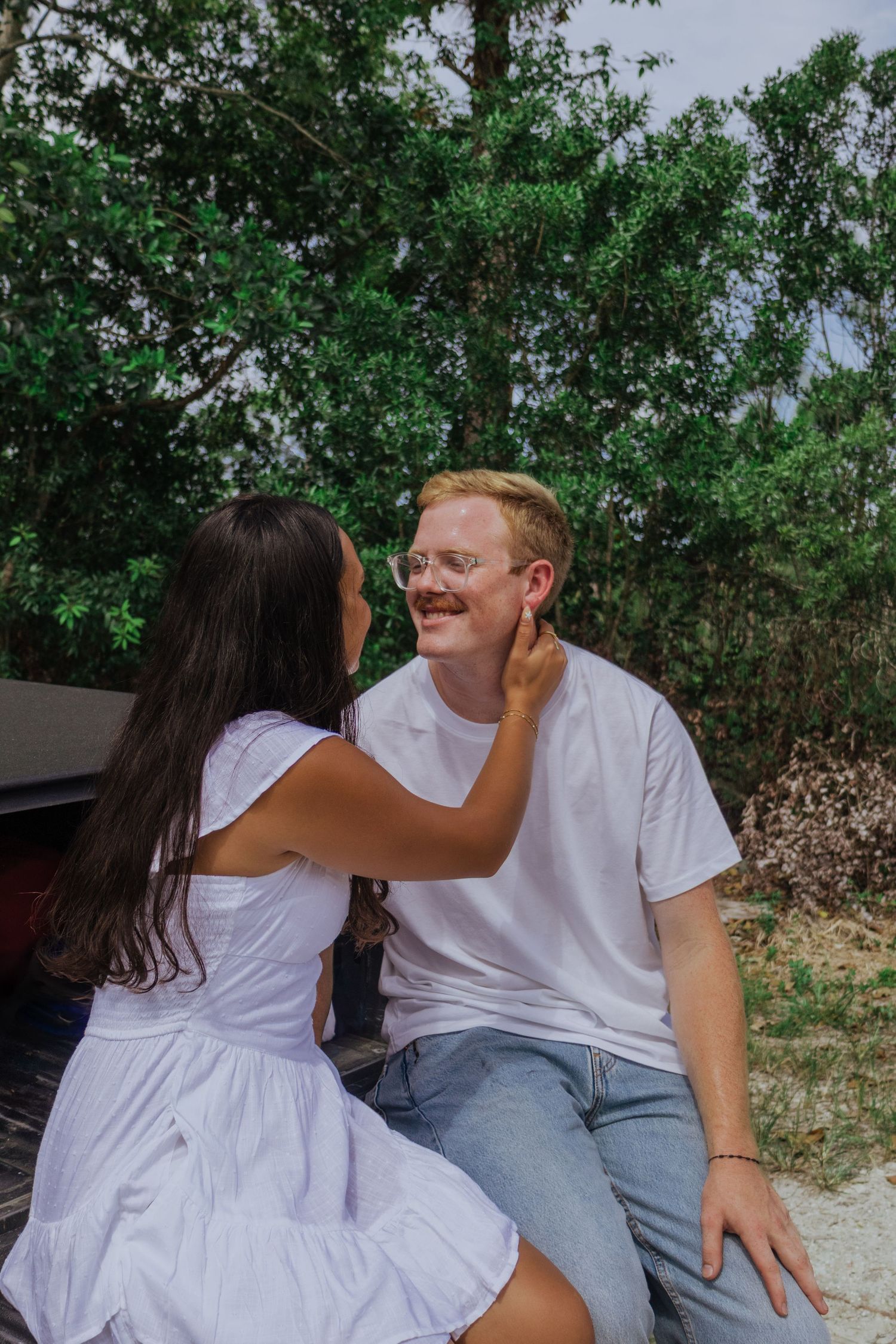 Smiling redhead man with glasses as his girlfriend gently touches his face outdoors.