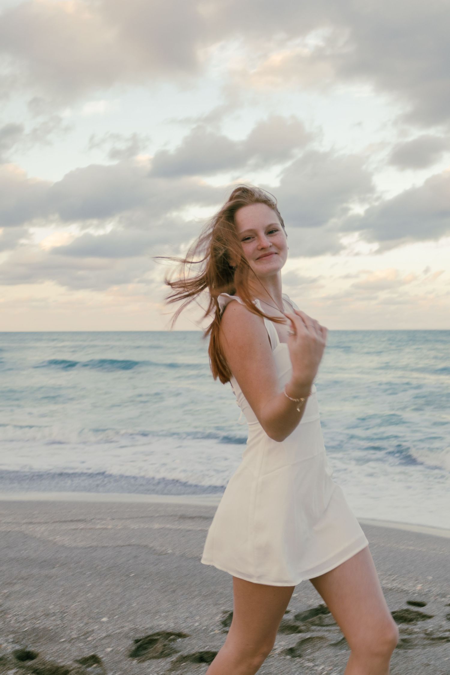 Smiling young woman in white mini dress with wind-blown hair posing on rocky beach at golden hour.