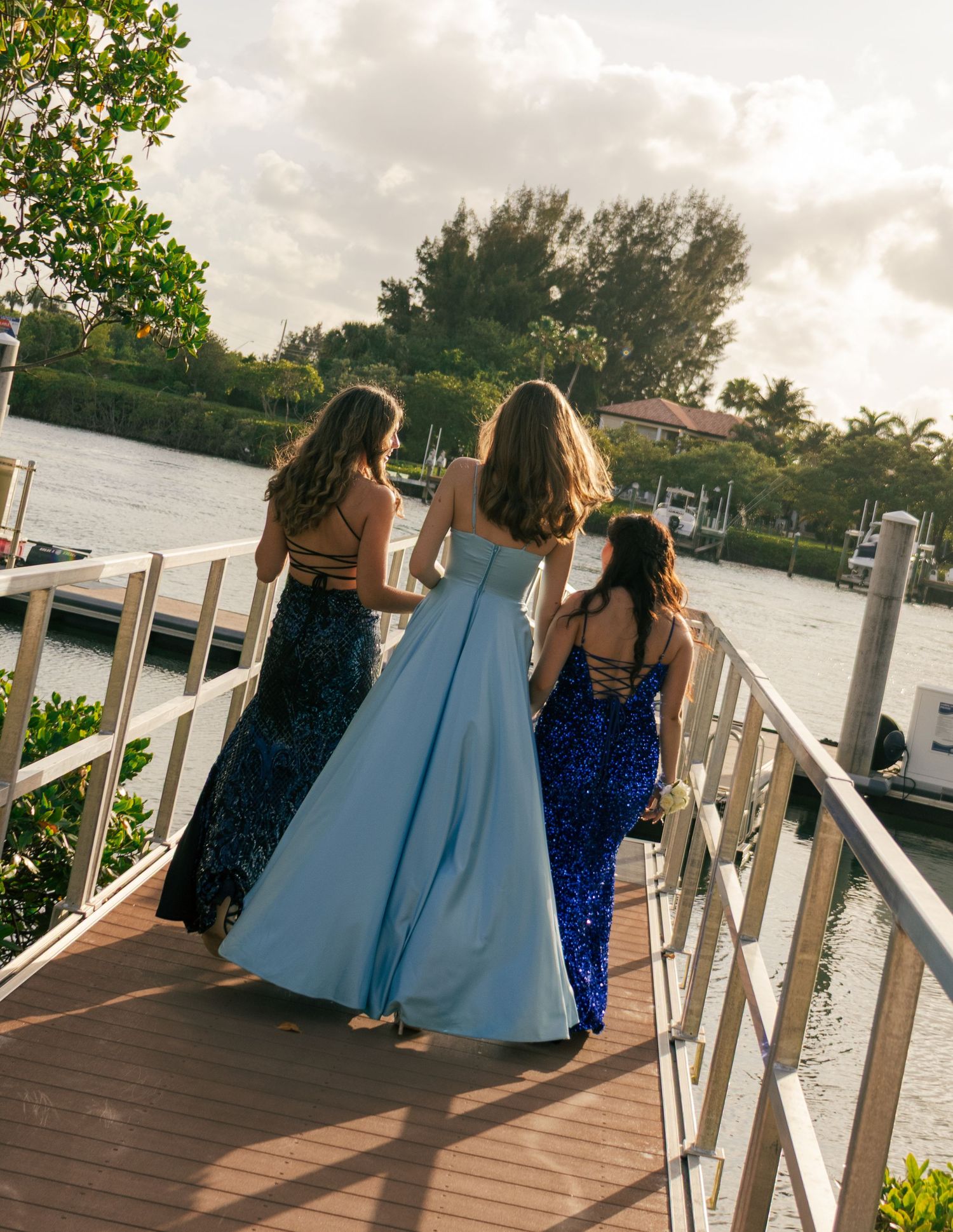 Three girls in formal prom gowns walk along a wooden dock overlooking a scenic Florida waterway.
