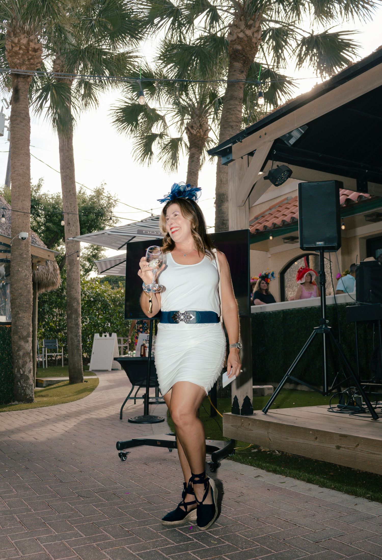 Woman in white dress holding a small dog near a outdoor stage with palm trees at a tropical venue.