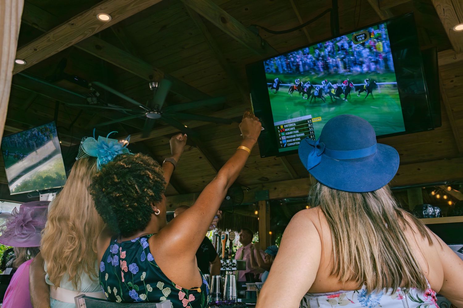 Two women in floral hats watching horse racing on TV screens at a bar.