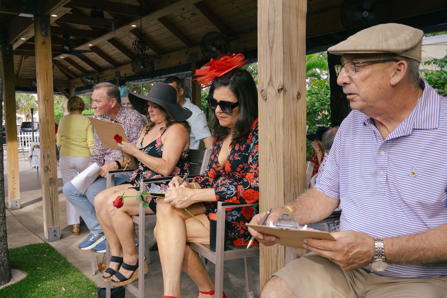 Guests in Derby hats sit under pavilion, reviewing programs at an outdoor Kentucky Derby watch party.