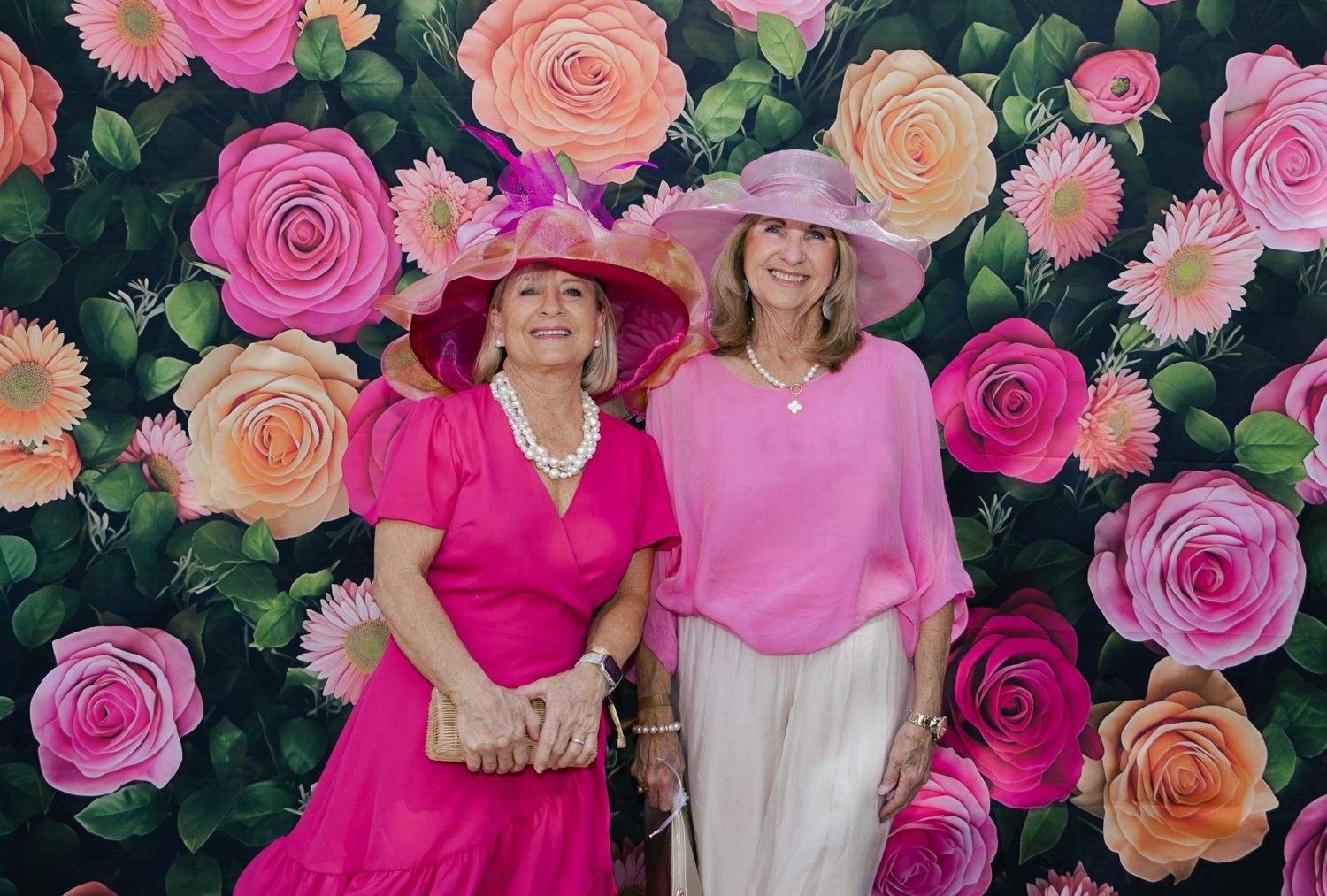 Two women wearing pink hats and outfits smile in front of a vibrant floral wall with roses and daisies.