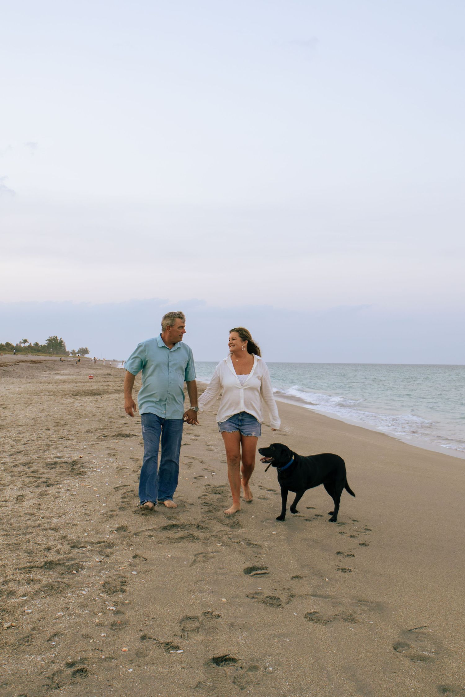 Middle-aged couple walking their black Labrador dog along a sandy beach at dusk.