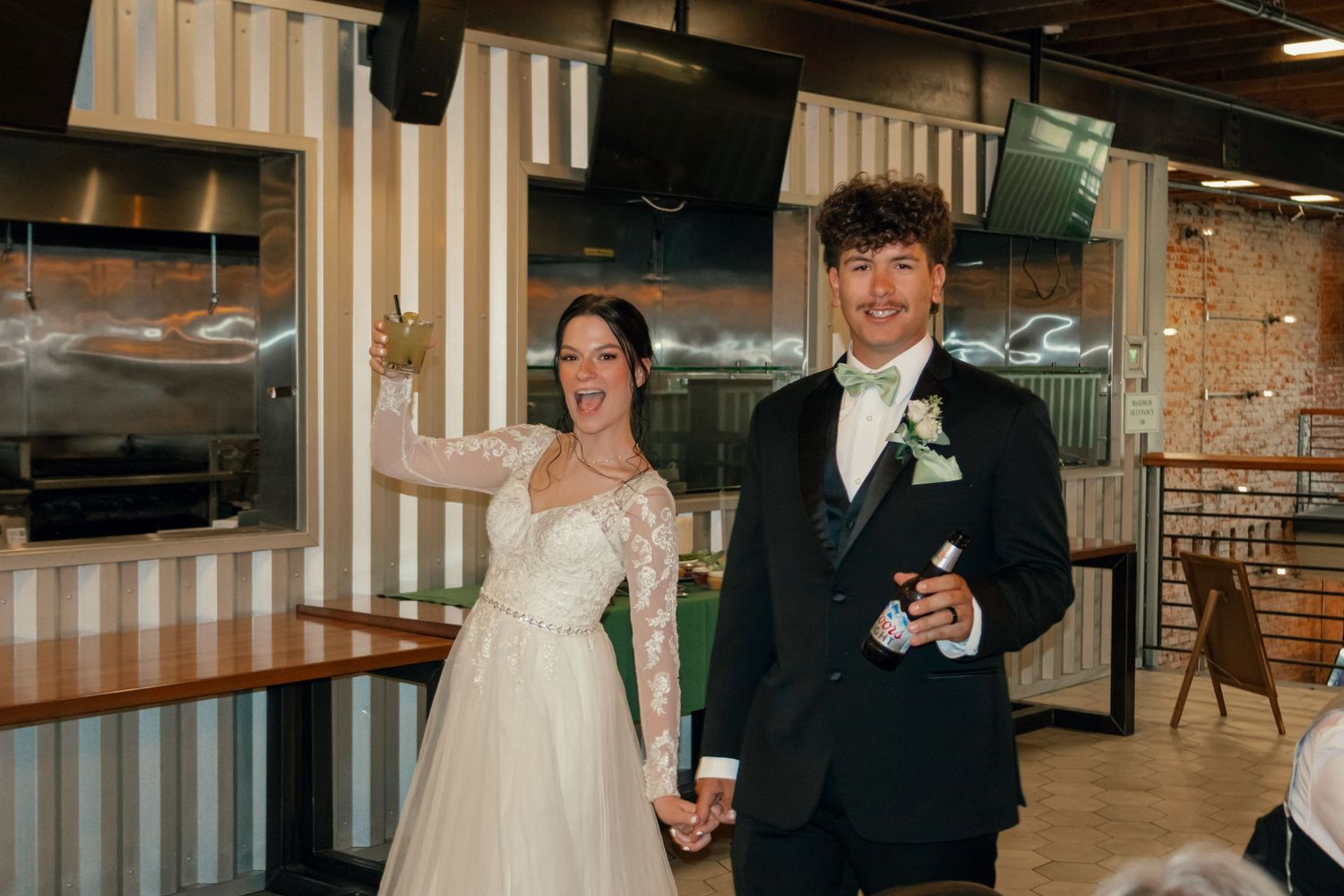 Joyful newlyweds hold hands and smile while entering their wedding reception venue with striped walls and neon signs.