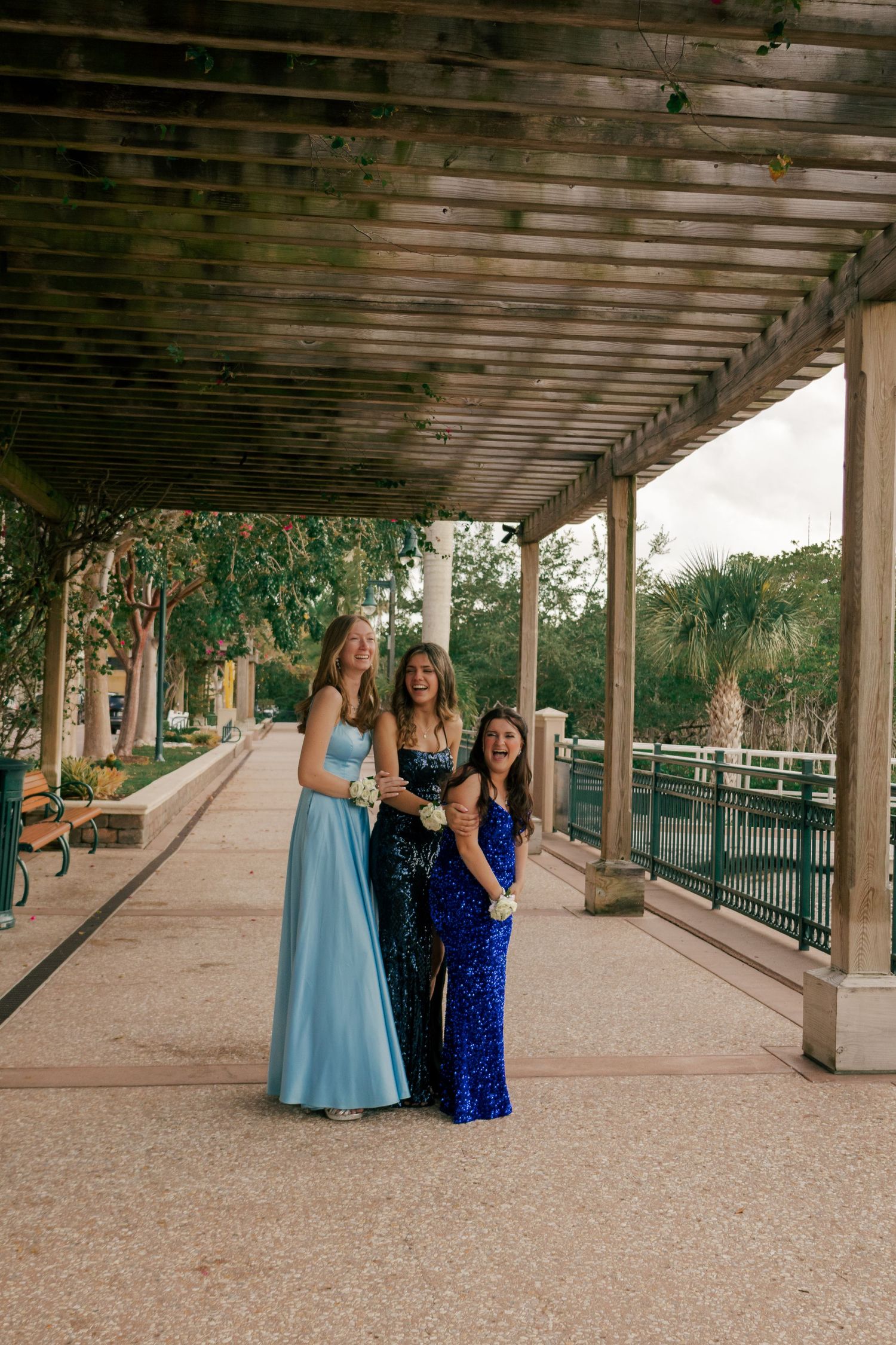 Three smiling teens in formal blue prom dresses stand along a scenic pergola-covered walkway.