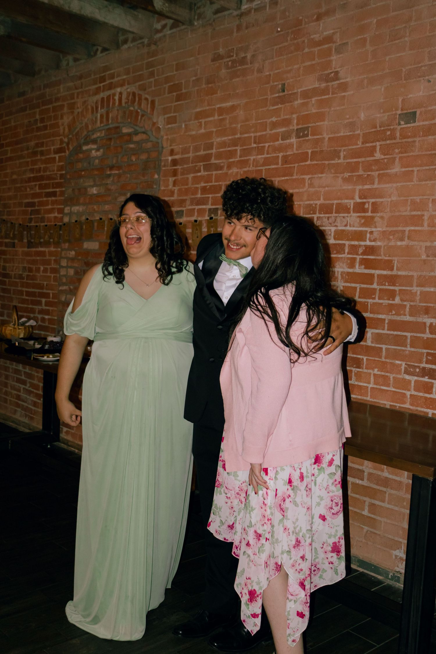 Three guests laugh together at wedding reception near exposed brick wall backdrop.