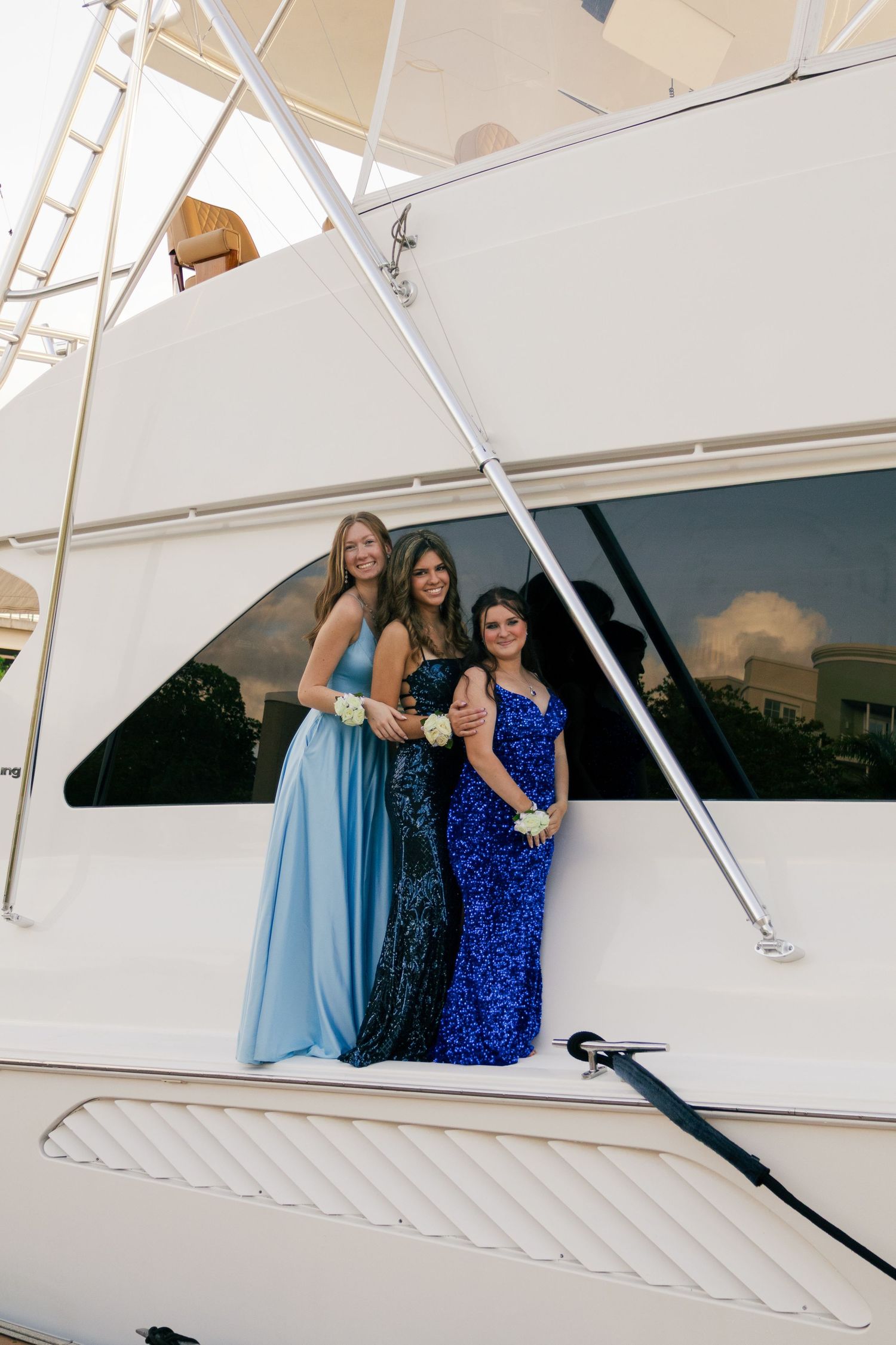 Three young women in blue sequin and satin prom gowns posing on white yacht.