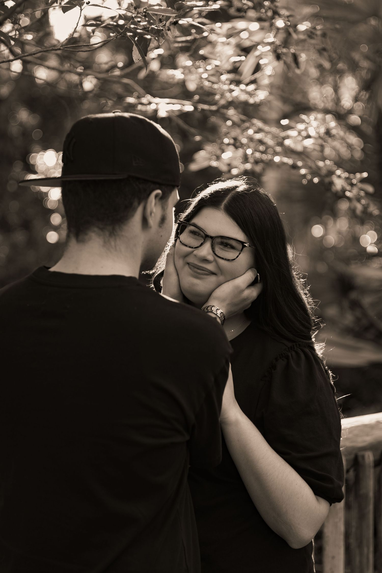 Sepia-toned photo of smiling woman with glasses gazing at man while he gently cups her face outdoors.