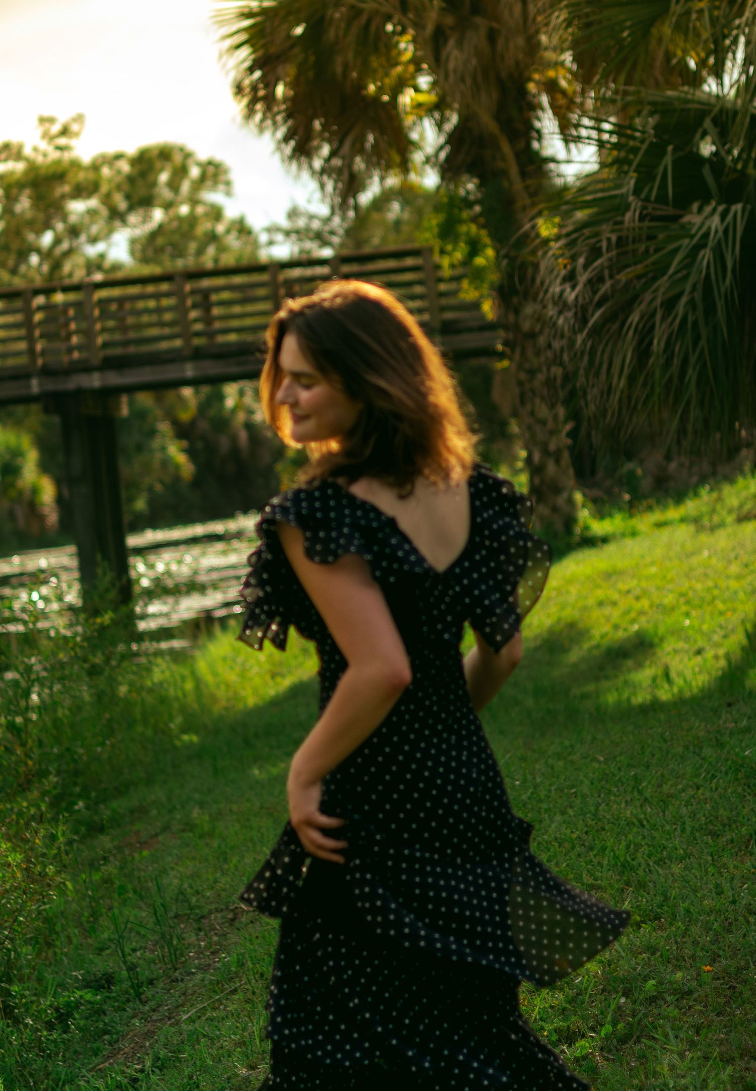 Woman in black polka dot dress twirling in sunlit park near wooden bridge and palm trees.
