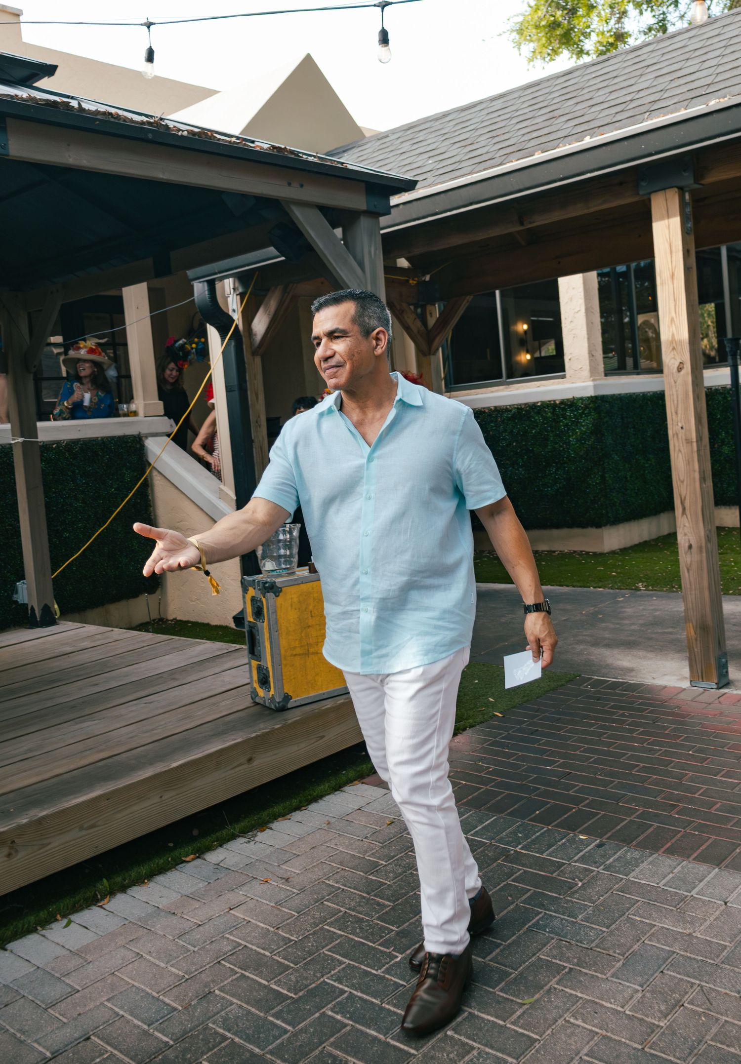Man in light blue shirt and white pants gestures at outdoor patio event.