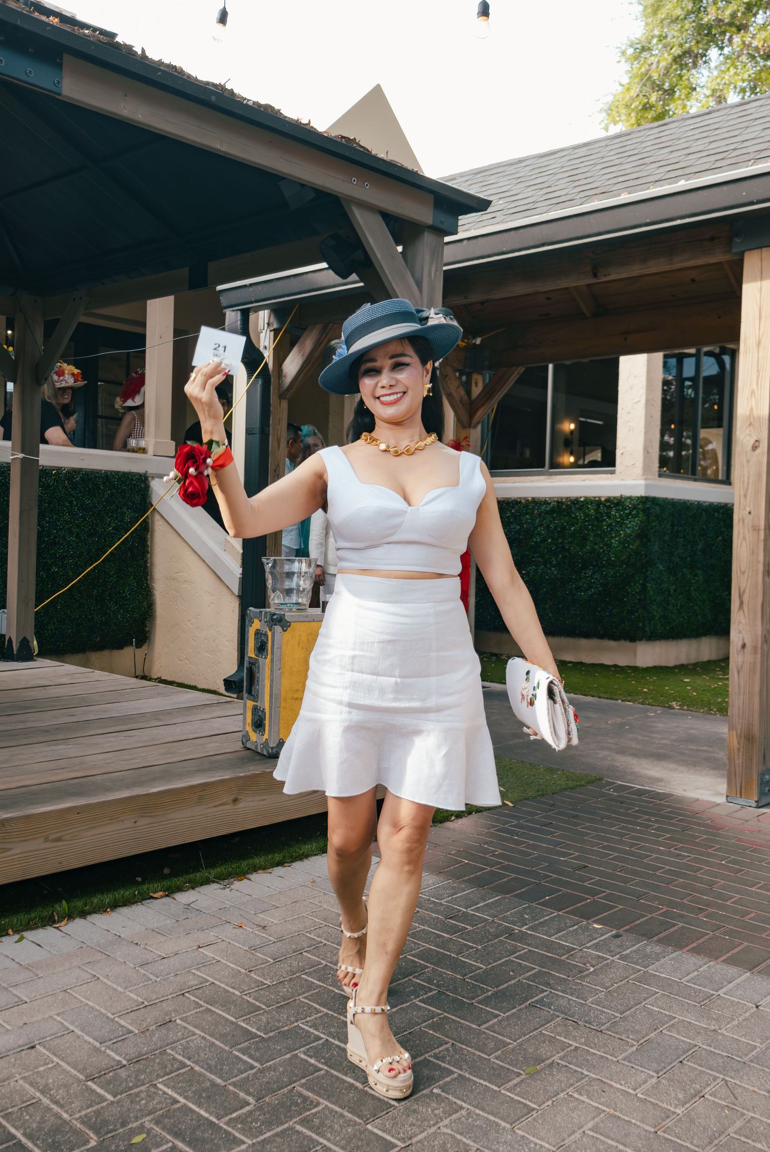 Woman in white two-piece outfit and navy hat holds card at outdoor Derby event.
