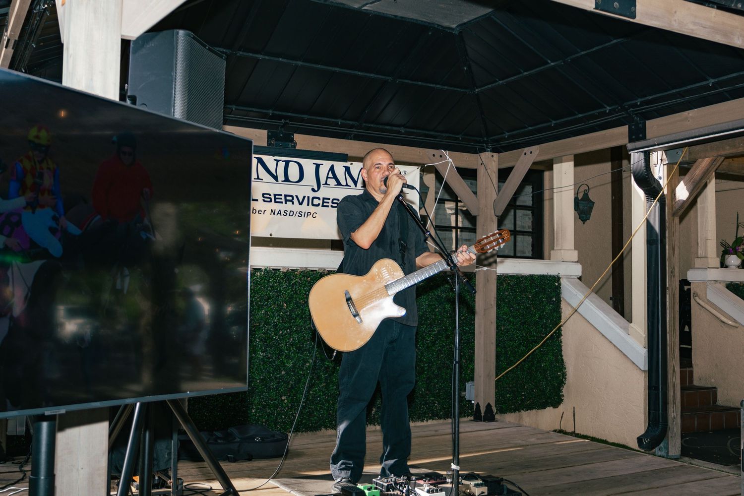 Musician performing on outdoor stage with acoustic guitar at a live event under a pavilion.