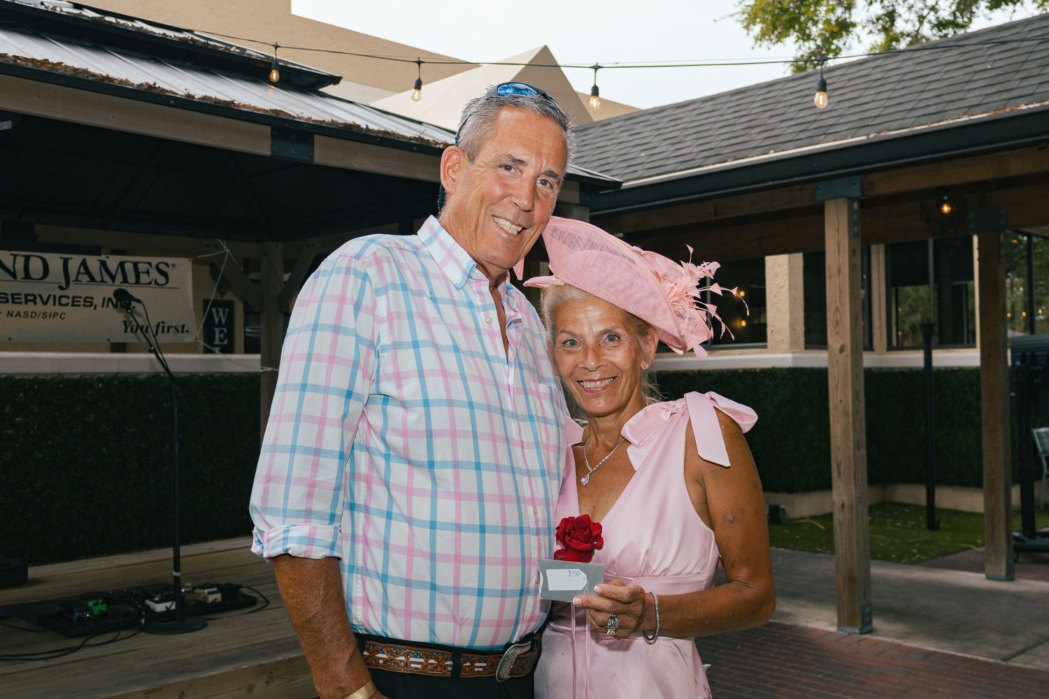 Smiling couple poses outdoors, woman wearing pink dress and decorative hat holding a rose.