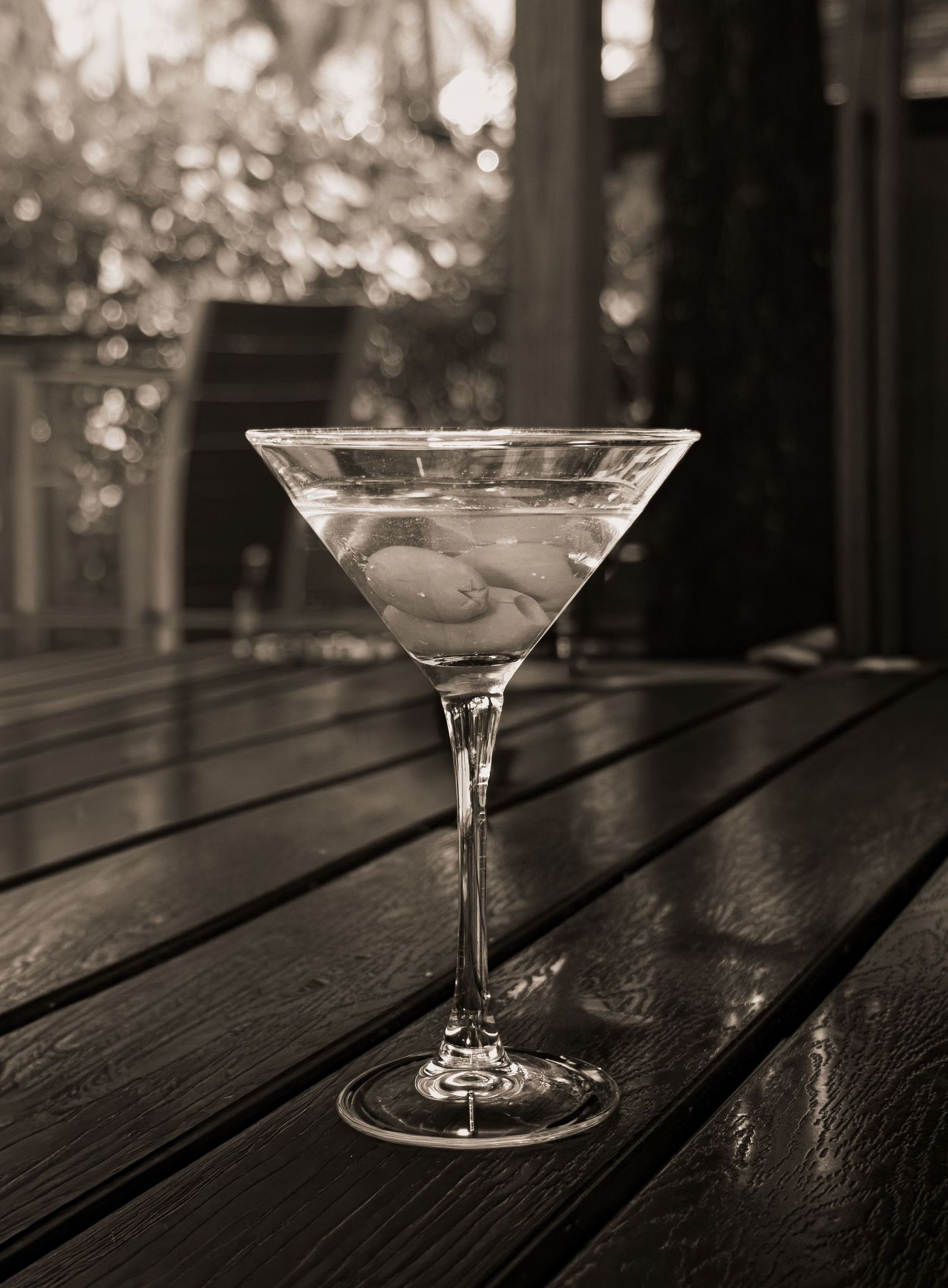 Classic dirty martini with olives in a crystal glass on a dark wooden deck in black and white.