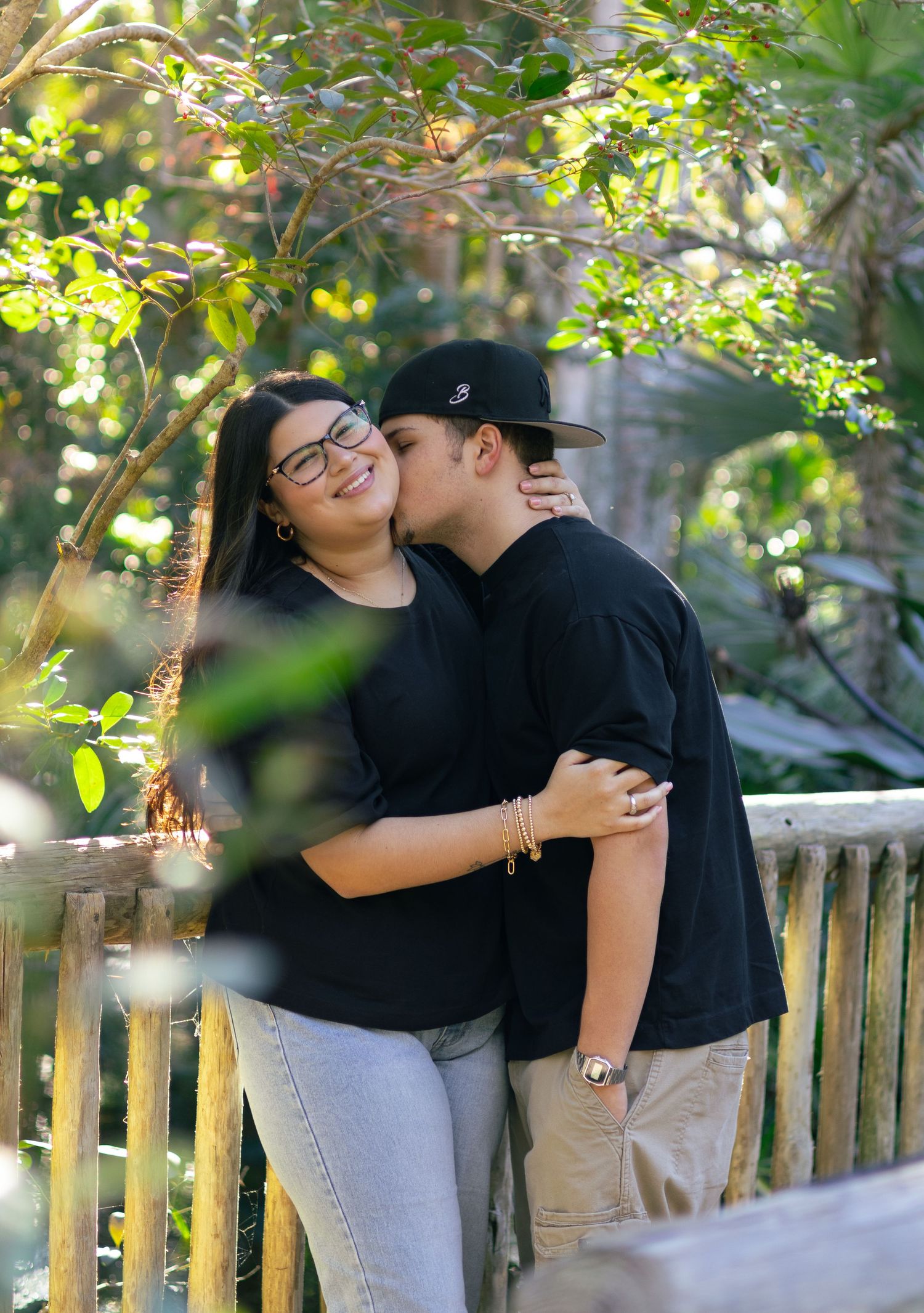 Young couple sharing a kiss outdoors on a wooden bridge surrounded by lush green trees.