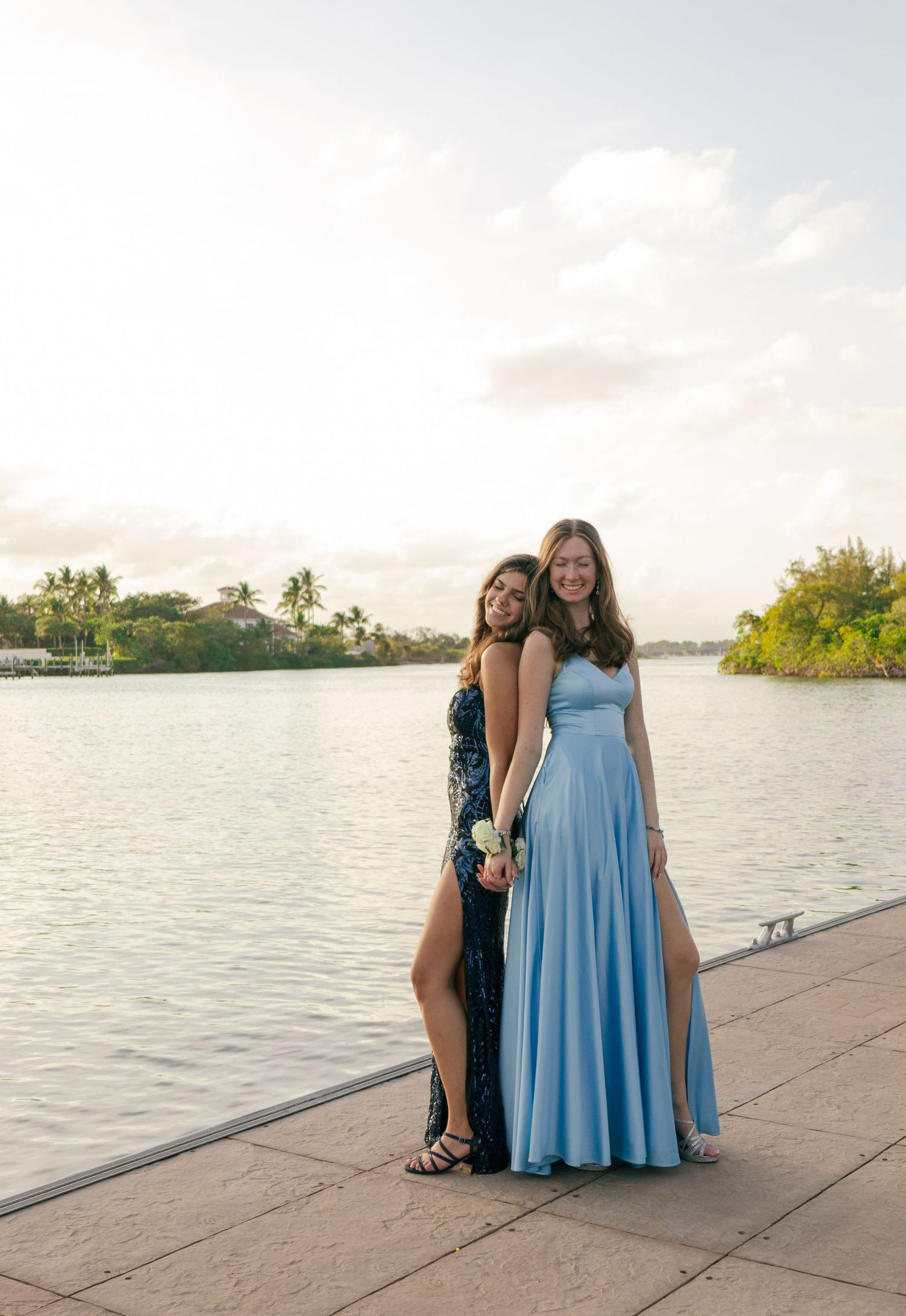 Two teenage girls in formal prom dresses pose together on a sunny waterfront promenade.