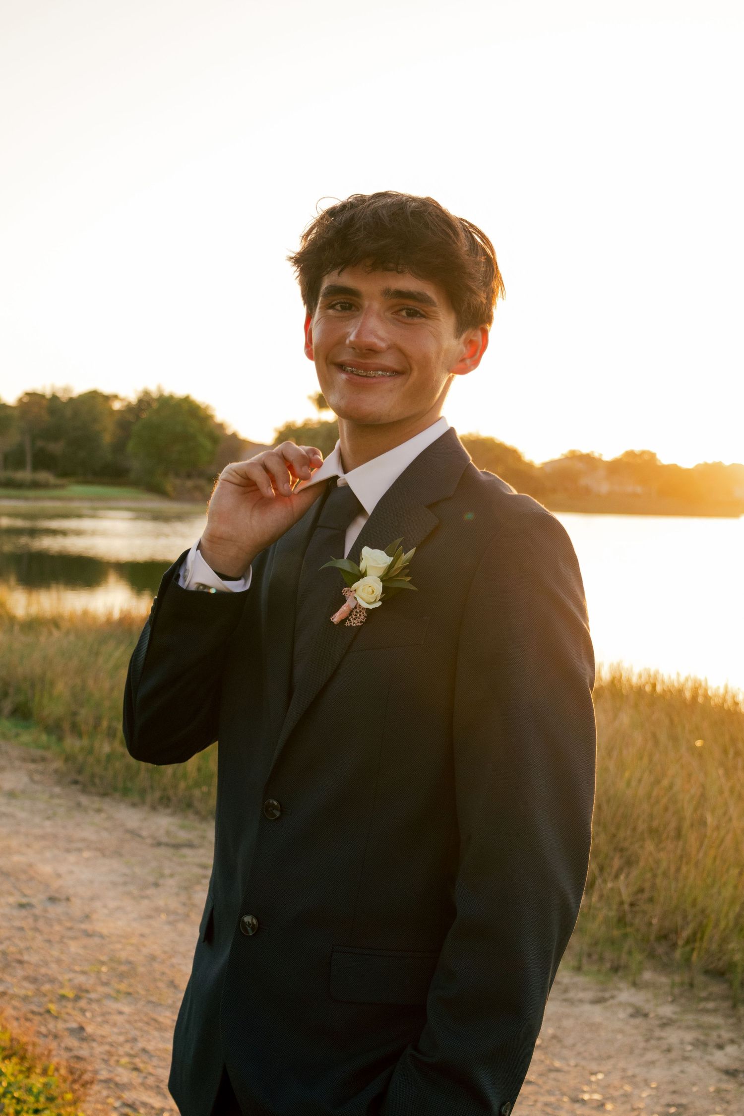 Smiling young man in black suit with boutonniere poses at golden sunset near lake.