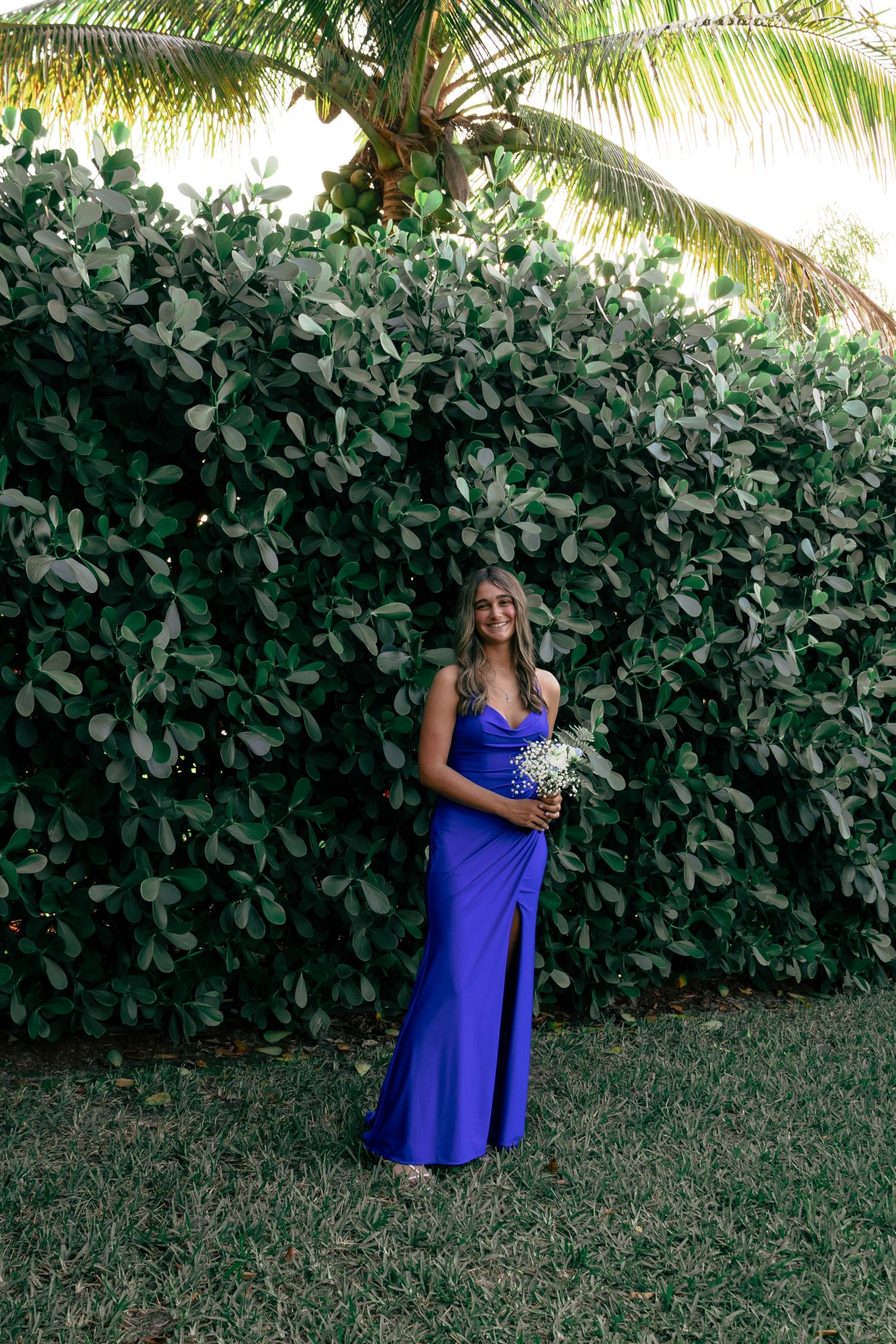 Young woman in a royal blue formal gown holding baby's breath bouquet by a green hedge.