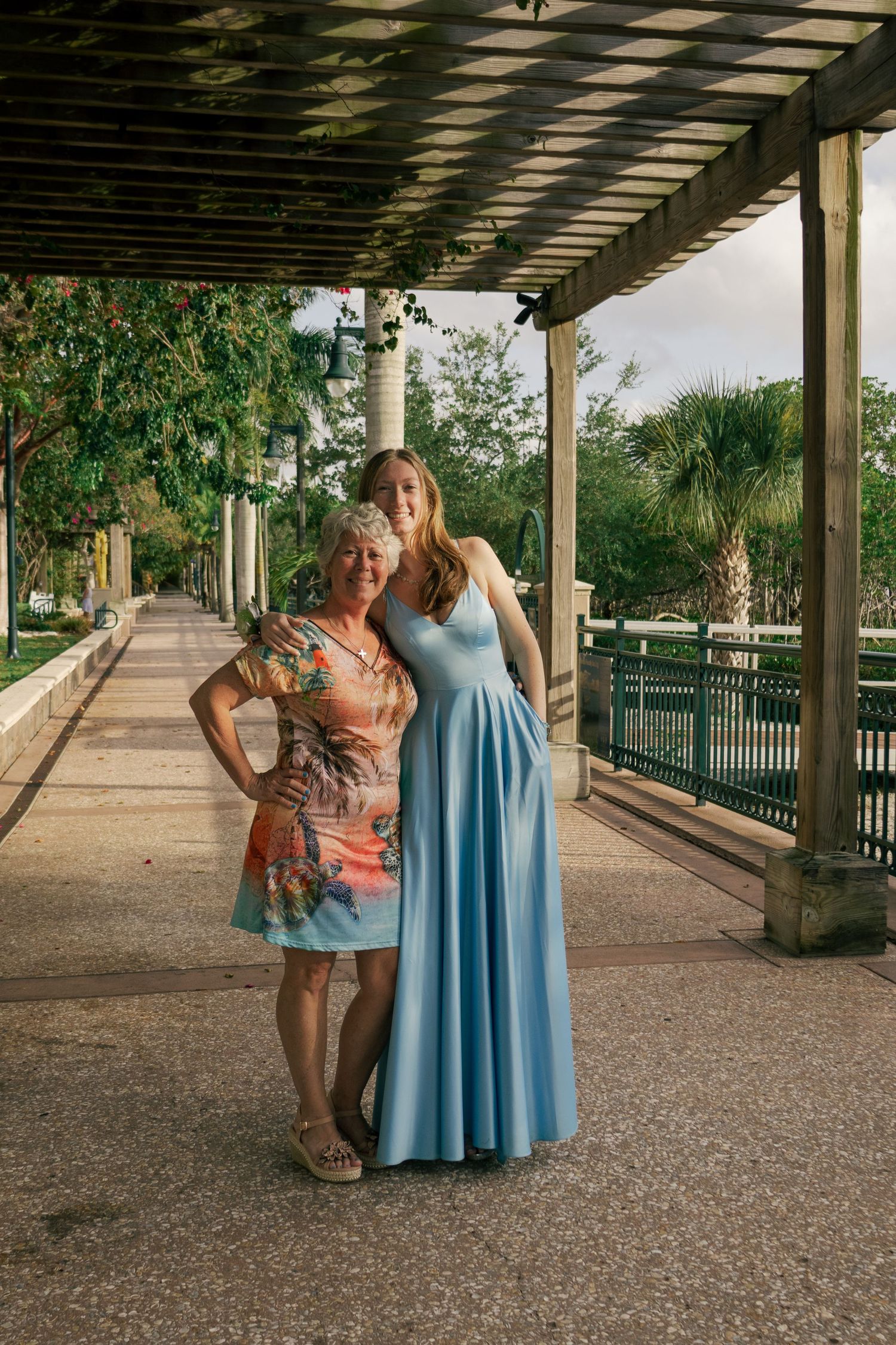 Young woman in light blue formal gown posing with older woman in floral dress under outdoor pergola.