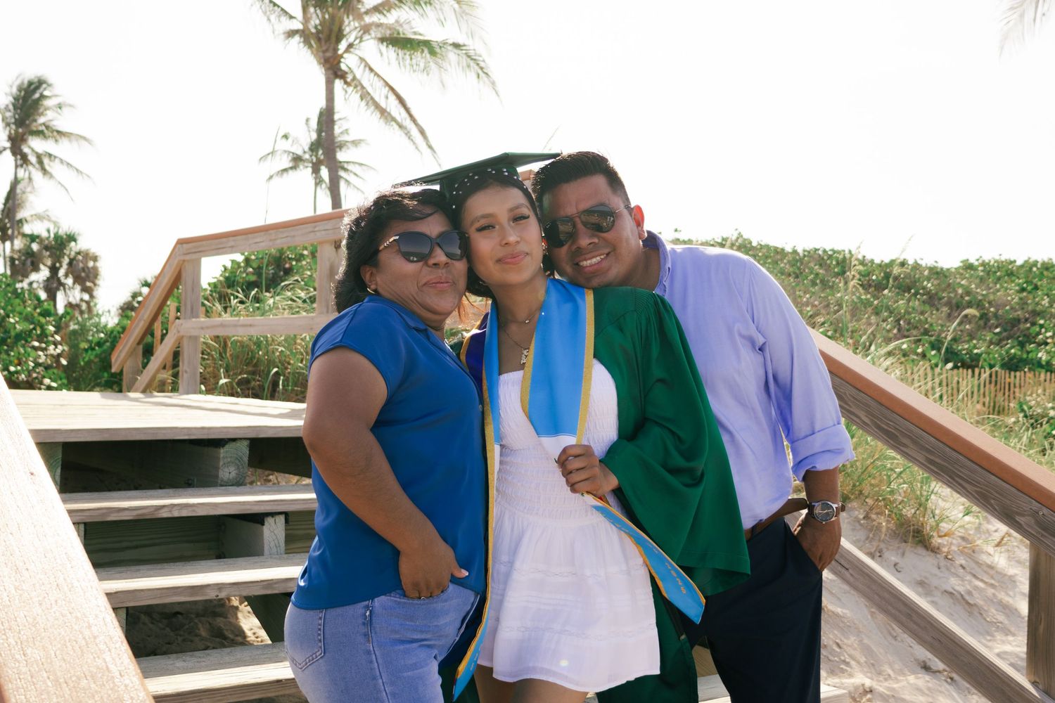 Three friends pose together on a beach boardwalk, smiling, with palm trees and sandy shore in the background.