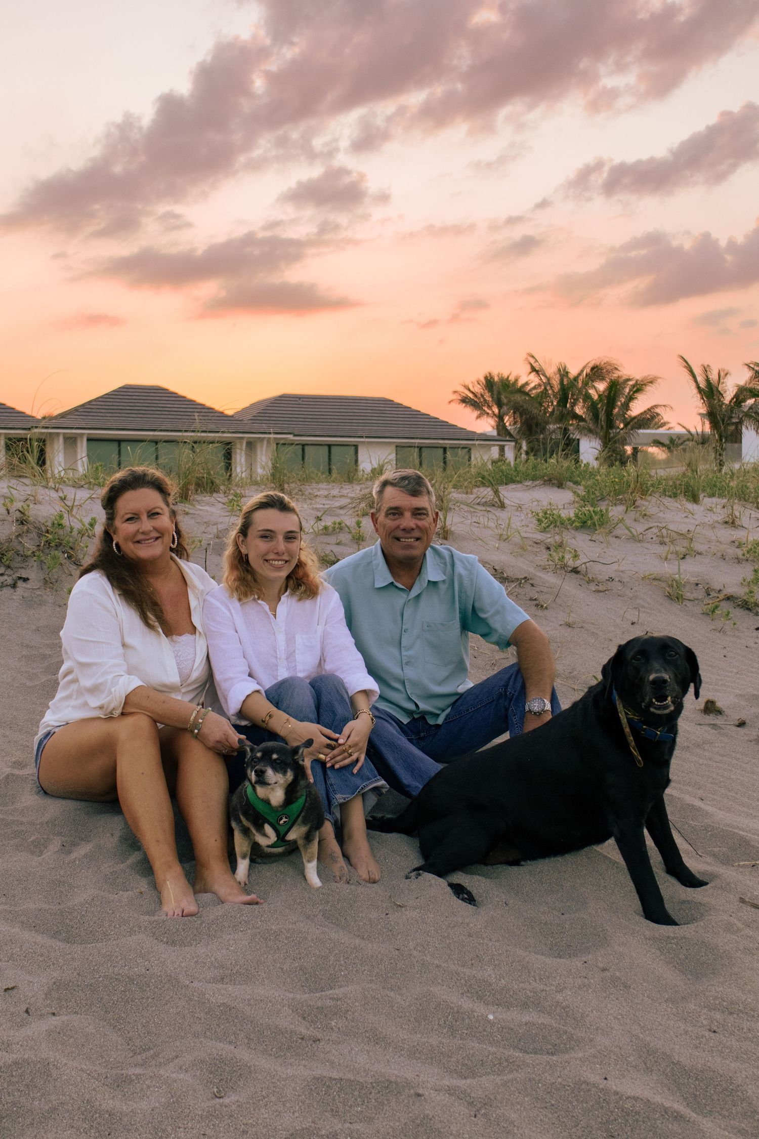 Family of three sitting on sandy beach at sunset with two dogs, tropical bungalows and palm trees in background.