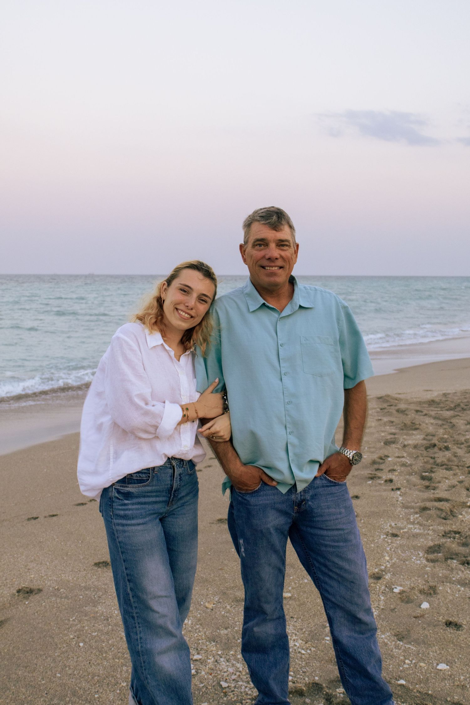 Smiling young woman and older man posing together on a sandy beach at twilight by the ocean.