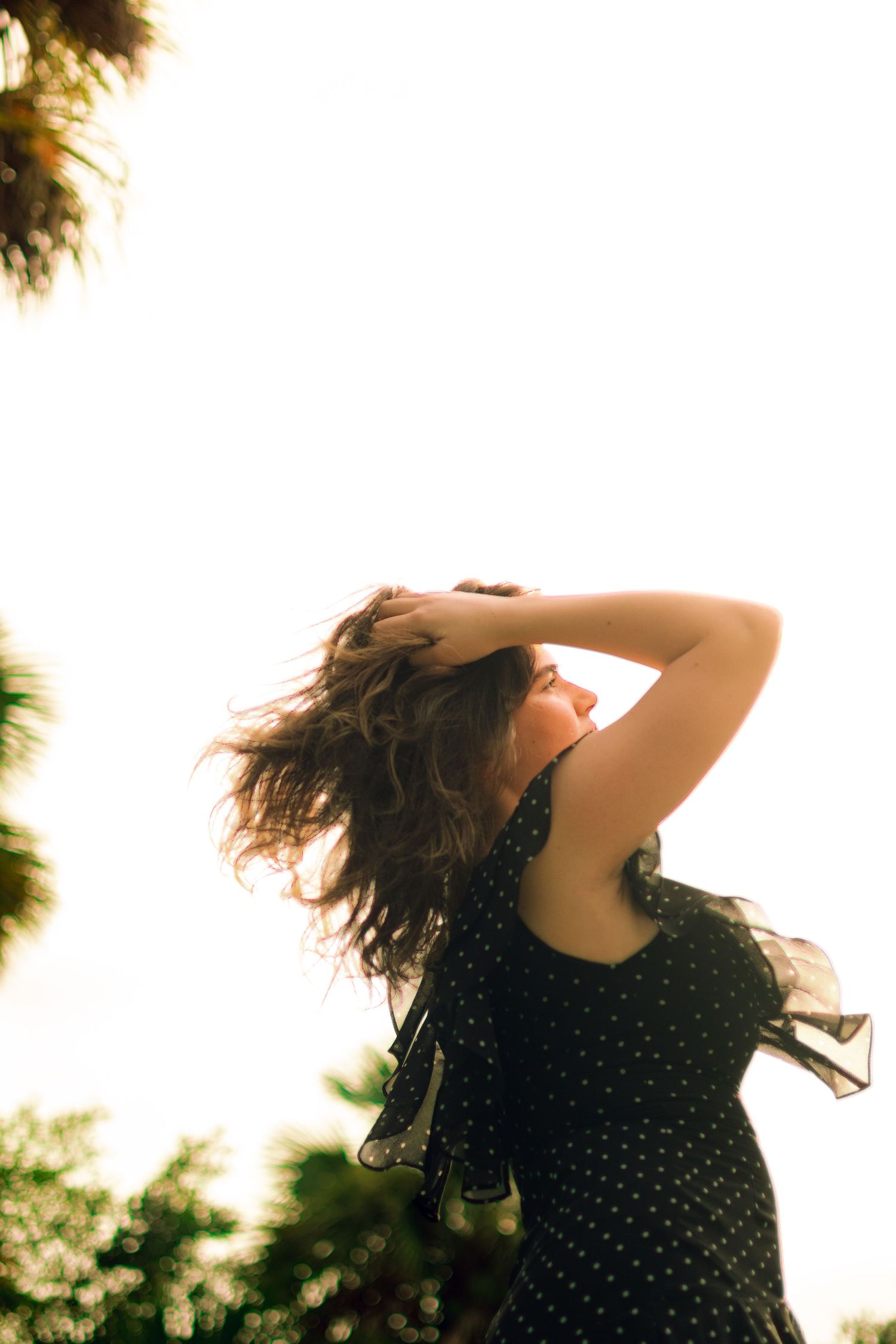 Woman in black polka dot dress holding hair back in breeze with bright sunlit sky background.